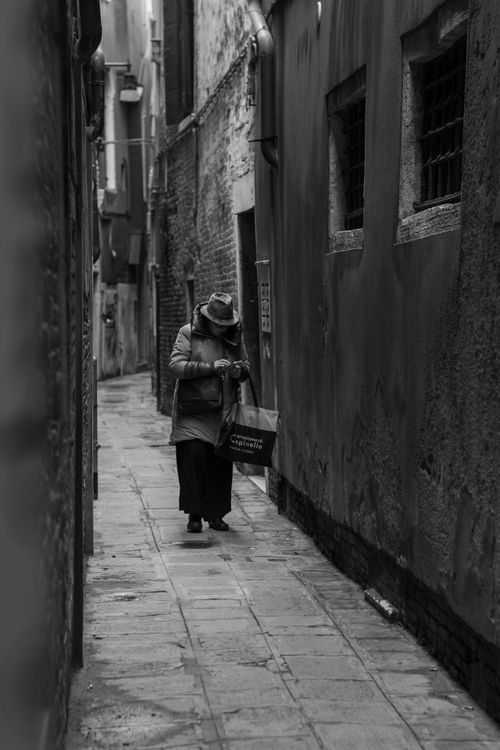Free A black and white image of an elderly woman walking alone in a narrow Venetian alley, showcasing the historic charm. Stock Photo