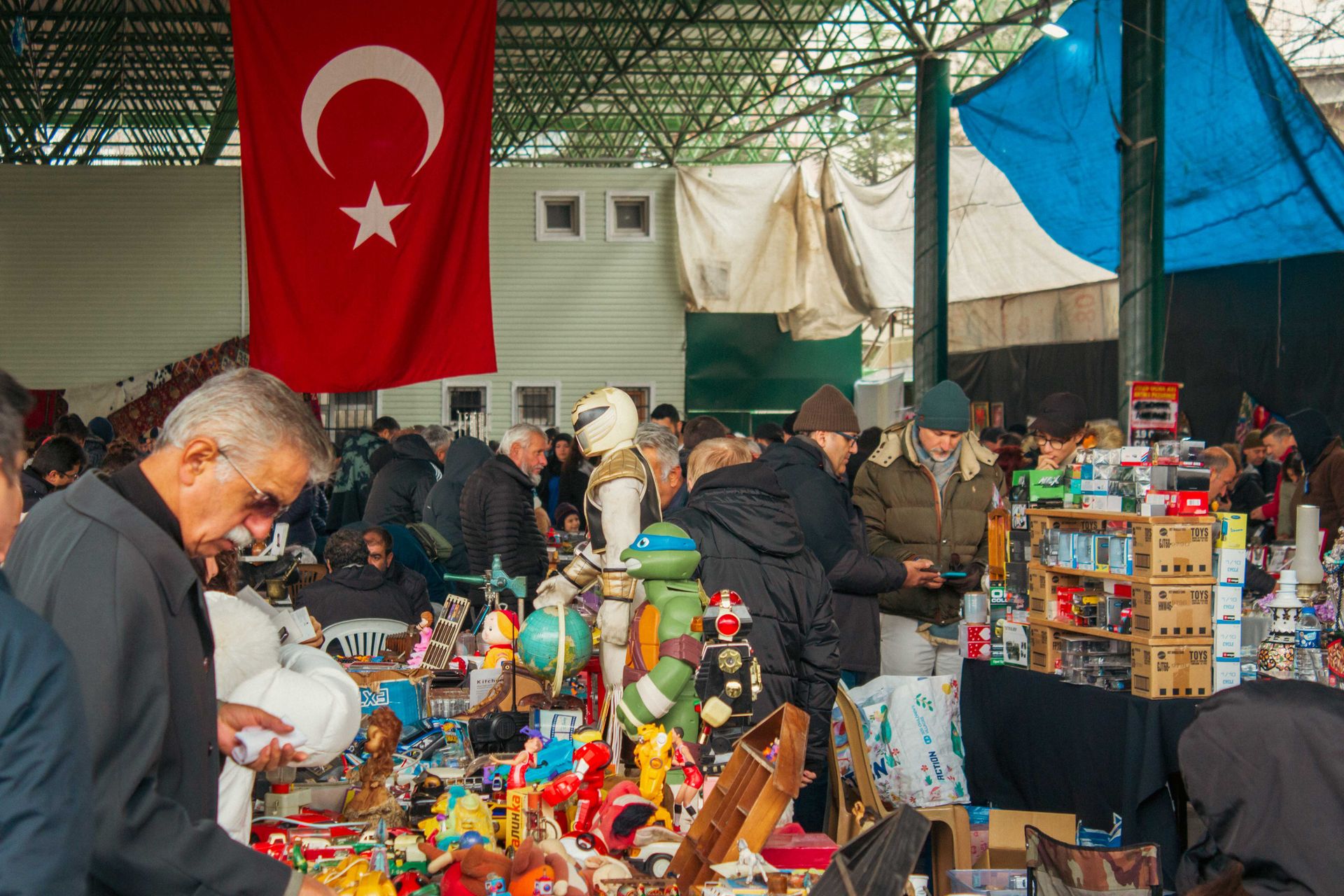 Busy flea market in Turkey with shoppers surrounded by antiques and toys under a Turkish flag.
