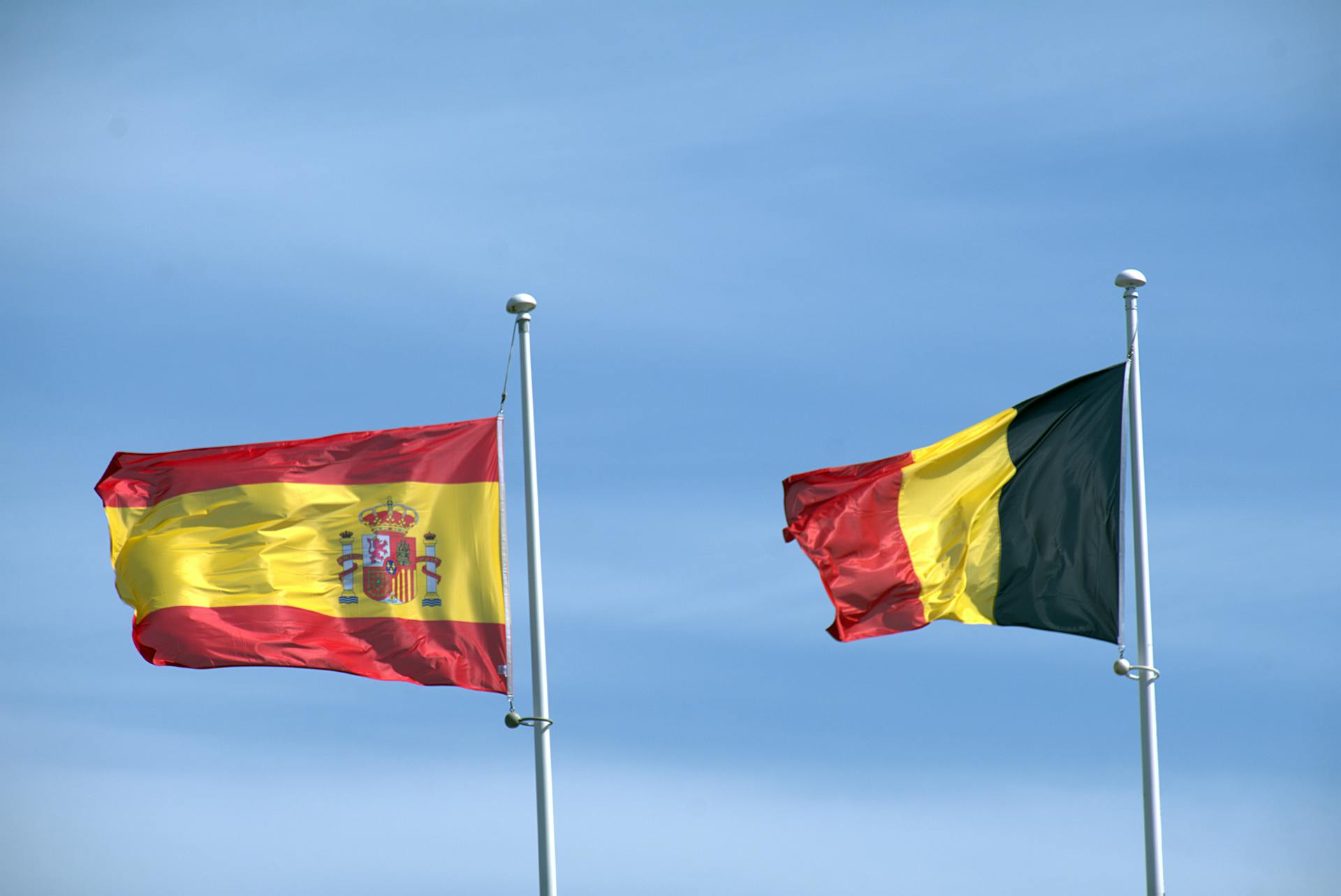 Flags of Spain and Belgium waving on poles under a blue sky, symbolizing international relations.