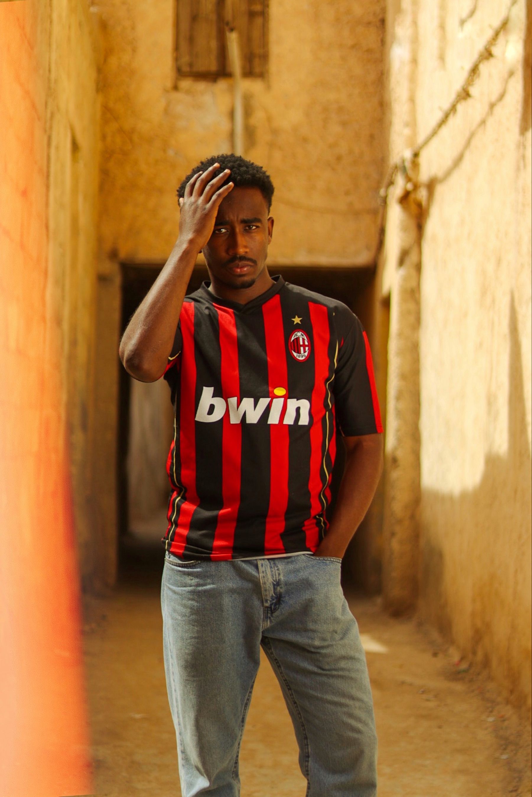 Young man in AC Milan jersey posing in a sunlit alleyway in Ouargla, Algeria.