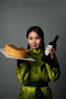 Smiling woman in green traditional attire holds a birthday cake and wine for celebration.
