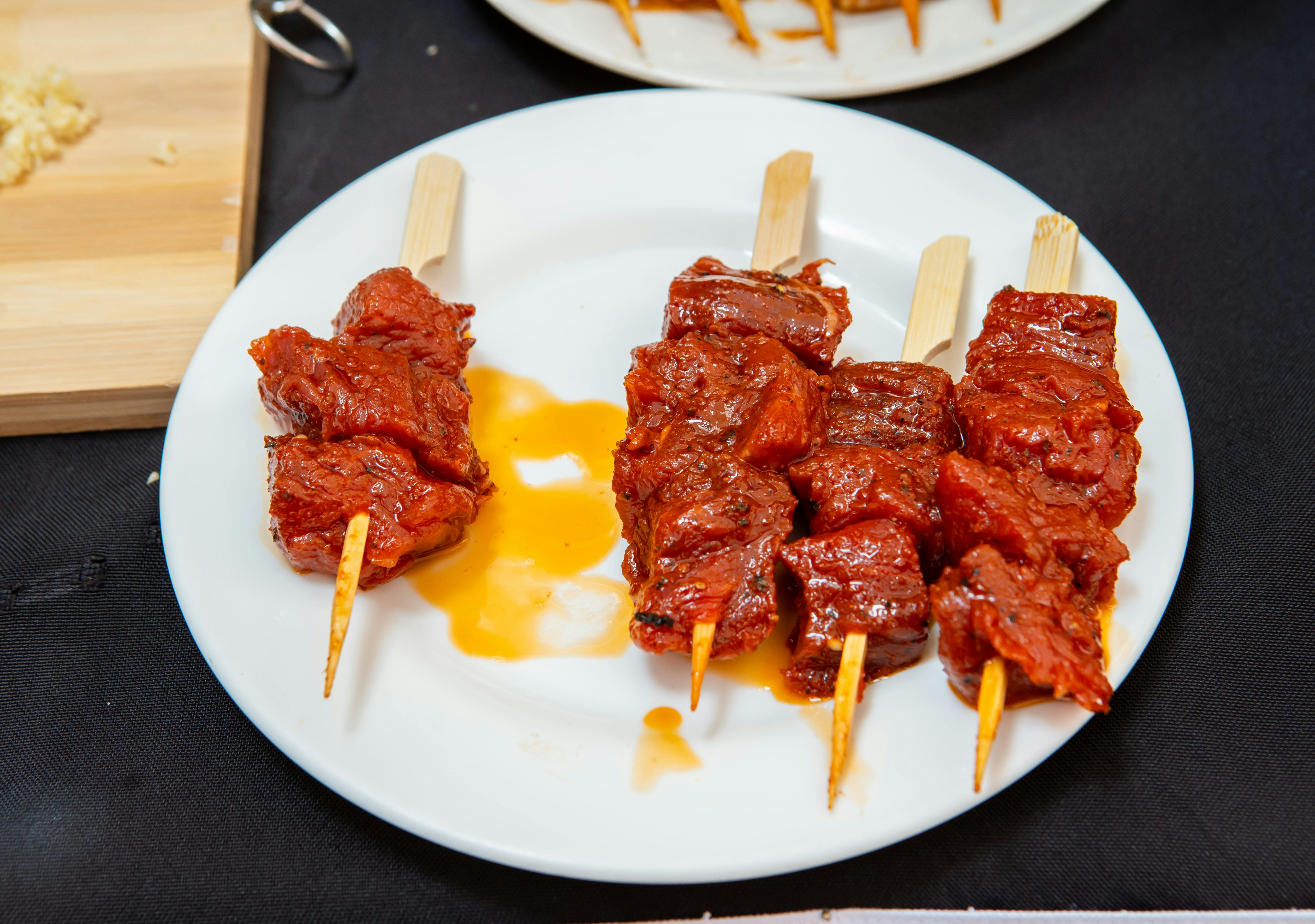 Grilled beef strips coated in reddish-brown suya spices on metal skewers, served alongside a small bowl of charred red pepper sauce on a white plate