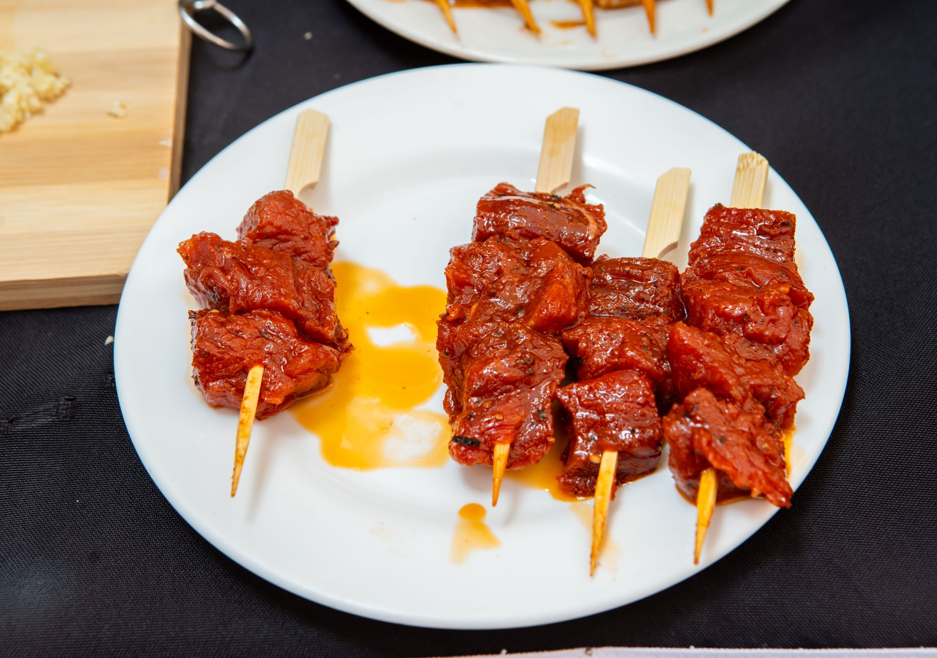 Grilled beef strips coated in reddish-brown suya spices on metal skewers, served alongside a small bowl of charred red pepper sauce on a white plate