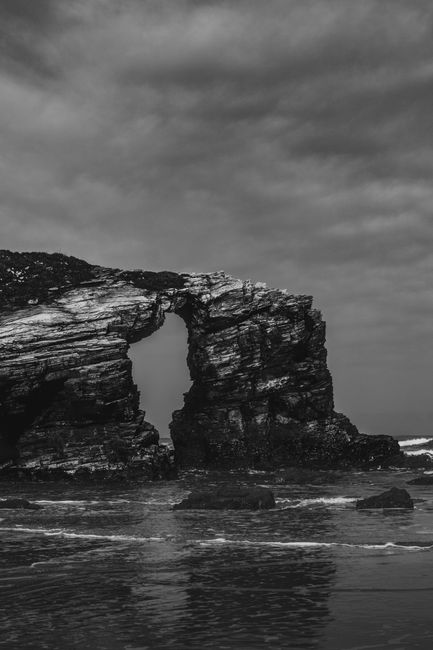 Moody black and white photo of a unique rock formation by the ocean shore with a cloudy sky.