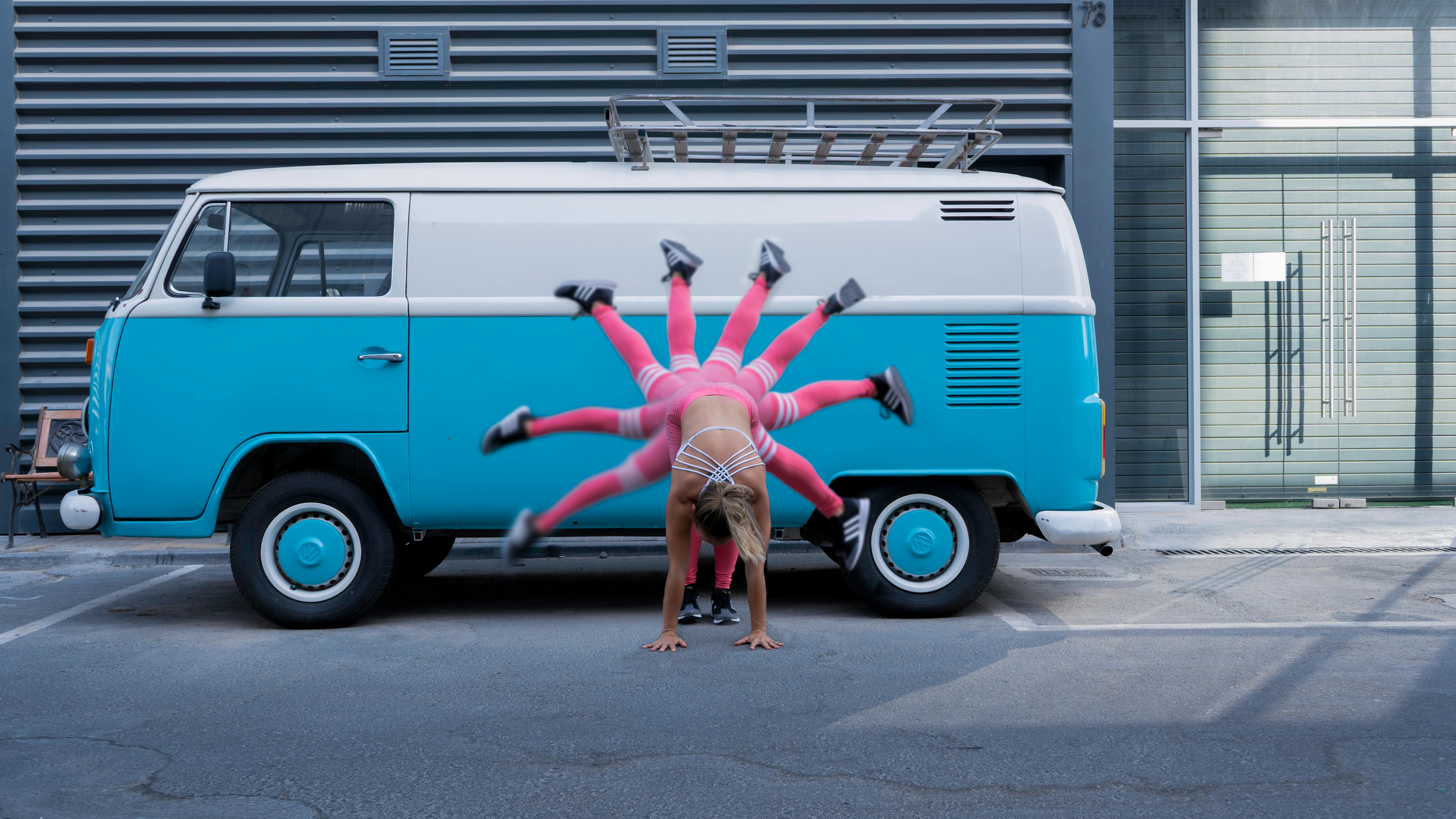 Woman doing an acrobatic handstand in front of a blue vintage van in an urban setting.