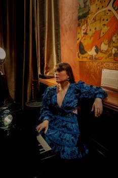 Elegant woman in a blue dress sits by a grand piano in a richly decorated room.