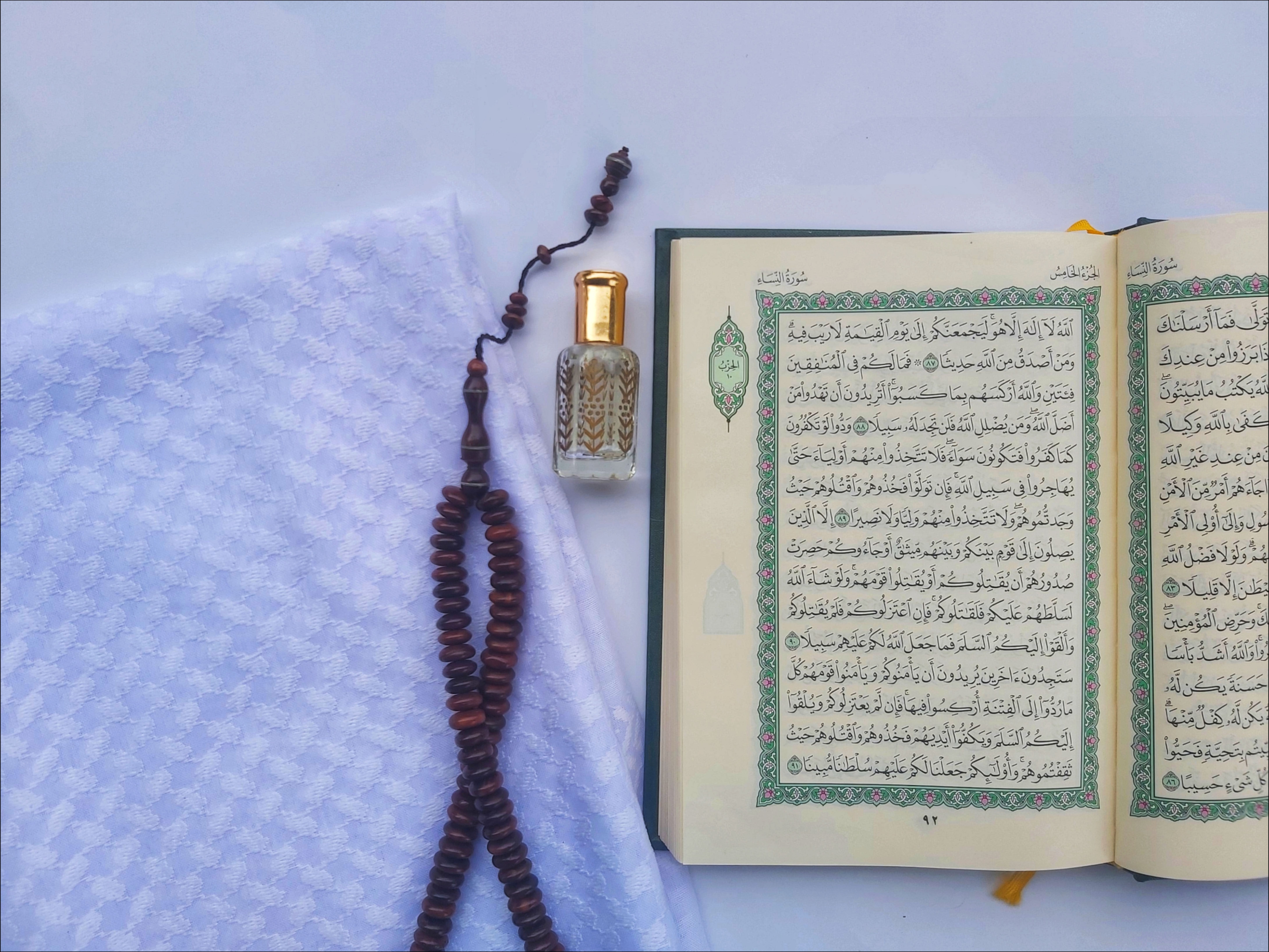 Open Quran with prayer beads and a perfume bottle on a white cloth background.