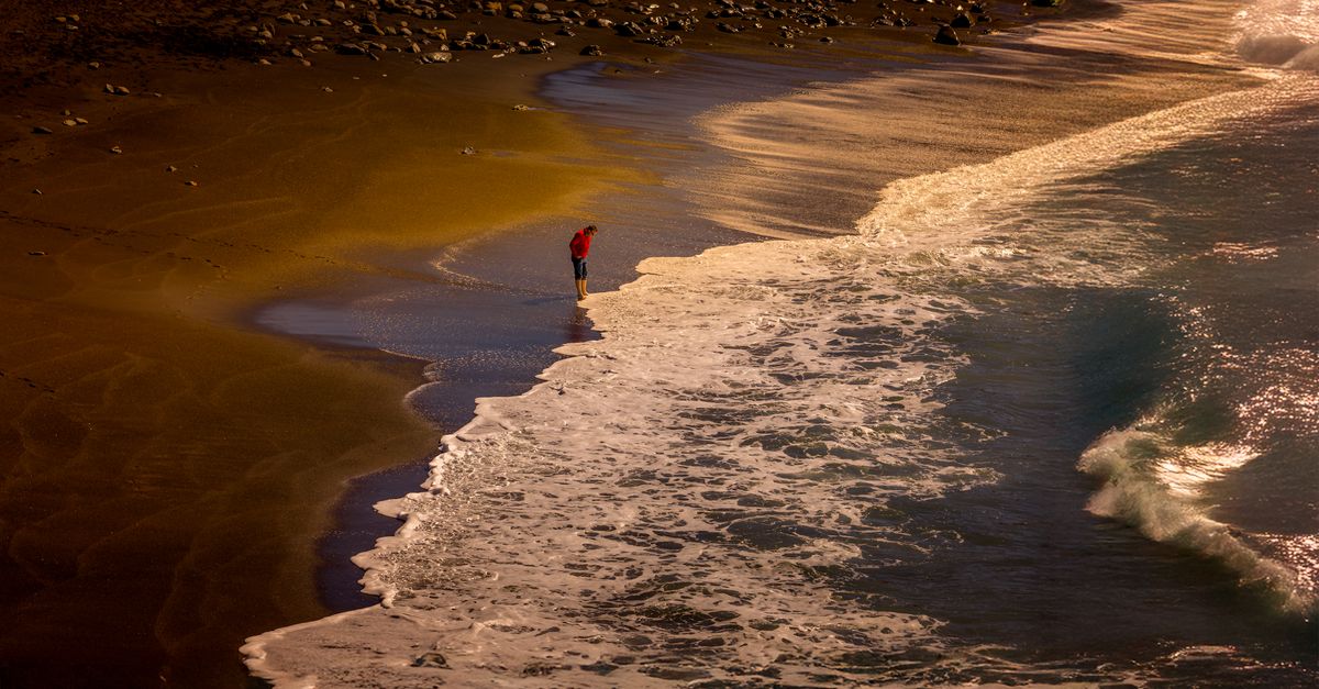 single person standing on a rocky shoreline at dusk looking at rough ocean waves, cinematic