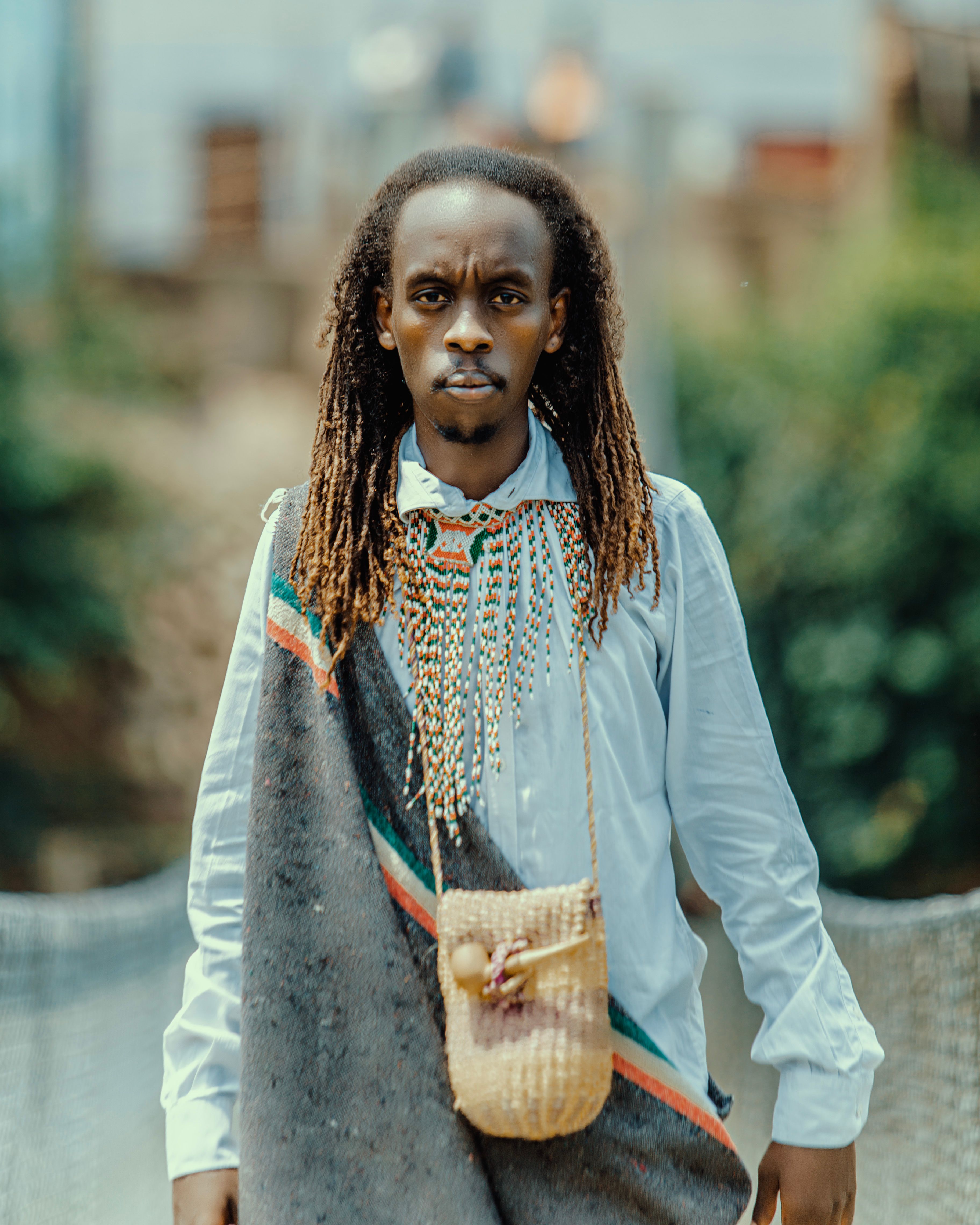 A man in traditional African attire with a woven bag in a lush outdoor setting.