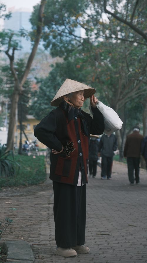 Free Elderly woman standing on a park path wearing Asian conical hat, holding a plastic bag. Stock Photo