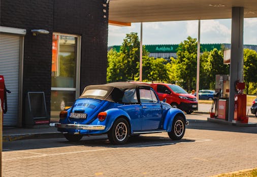 Blue vintage Volkswagen Beetle parked at modern gas station on a sunny day.