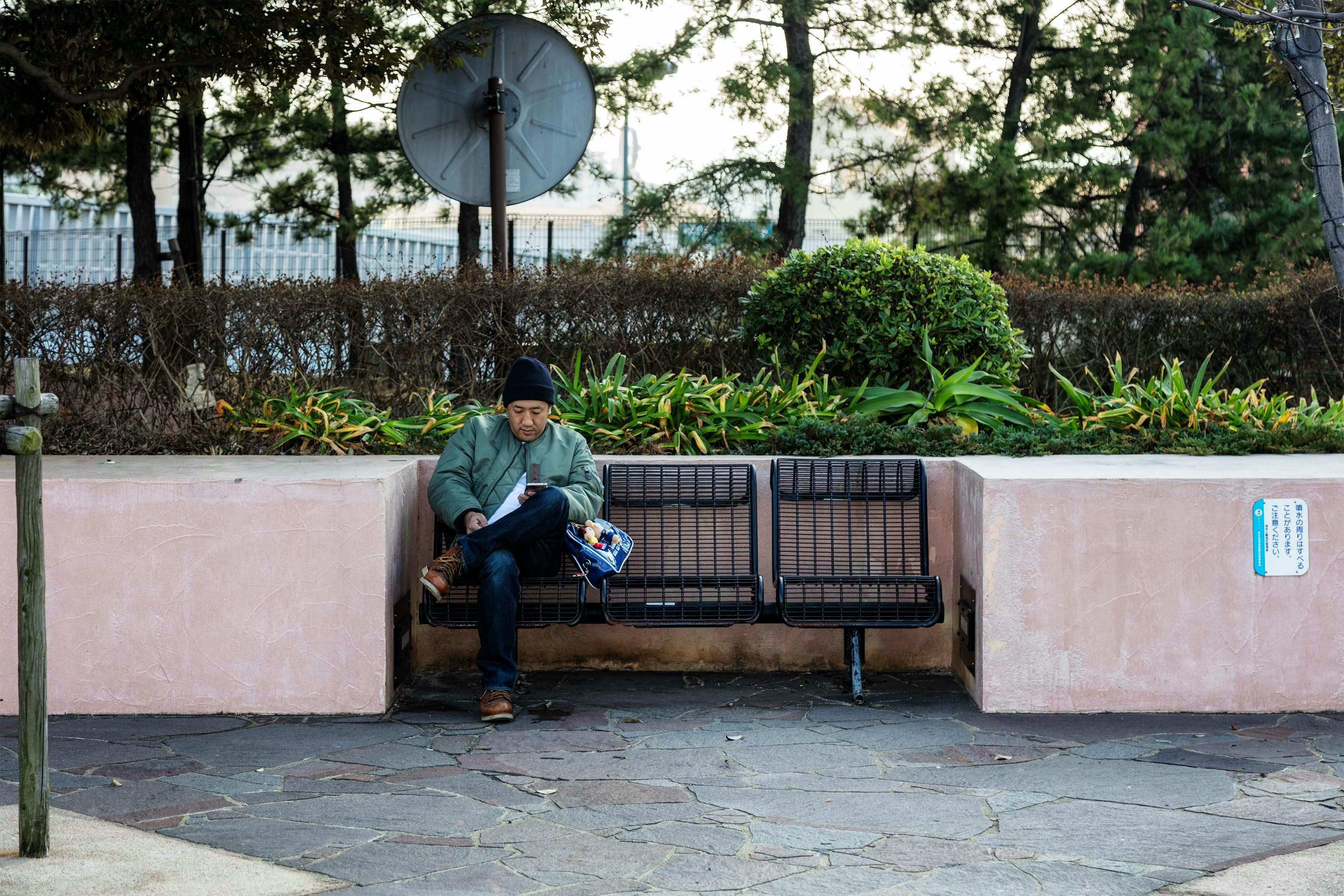 一名男子坐在公园的长凳上，周围绿植环绕，正全神贯注地玩着智能手机