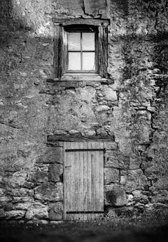 Black and white image of a rustic stone wall with a wooden door and window, vintage style.