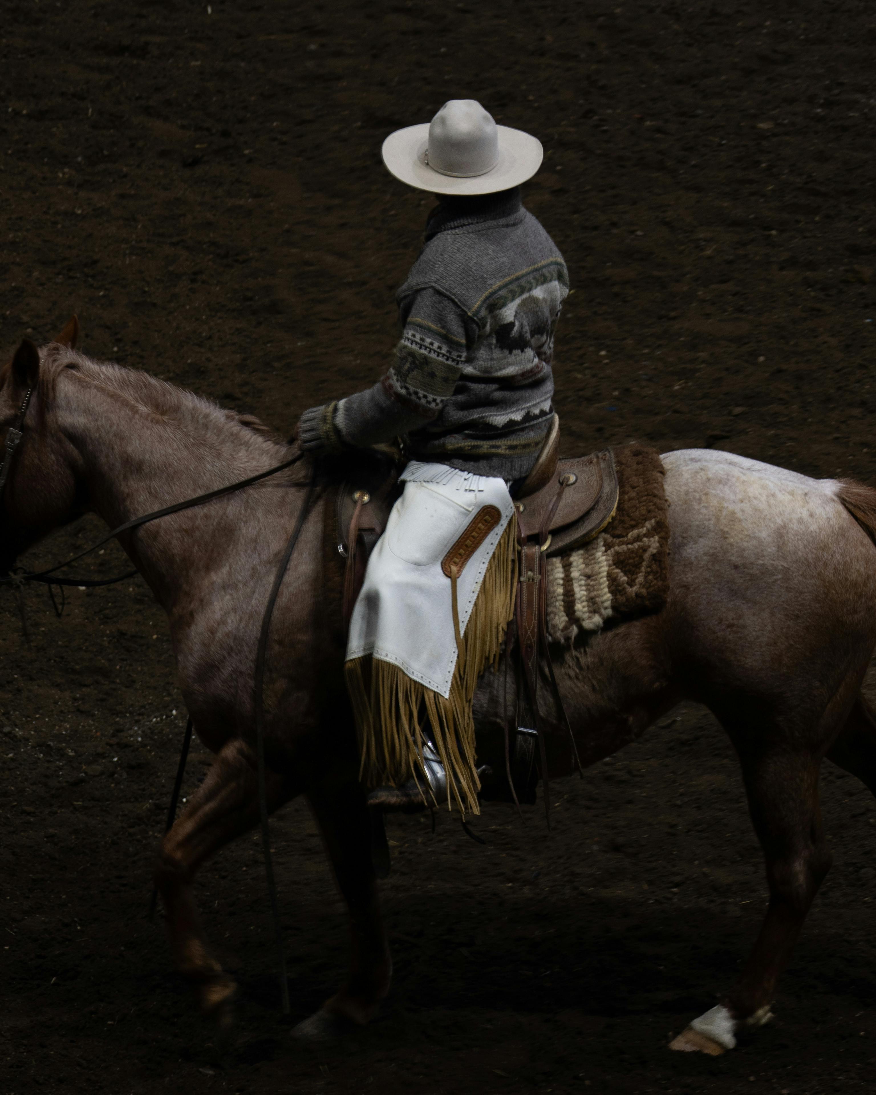 Western cowboy riding a horse with fancy saddle and fringed chaps in an outdoor setting.