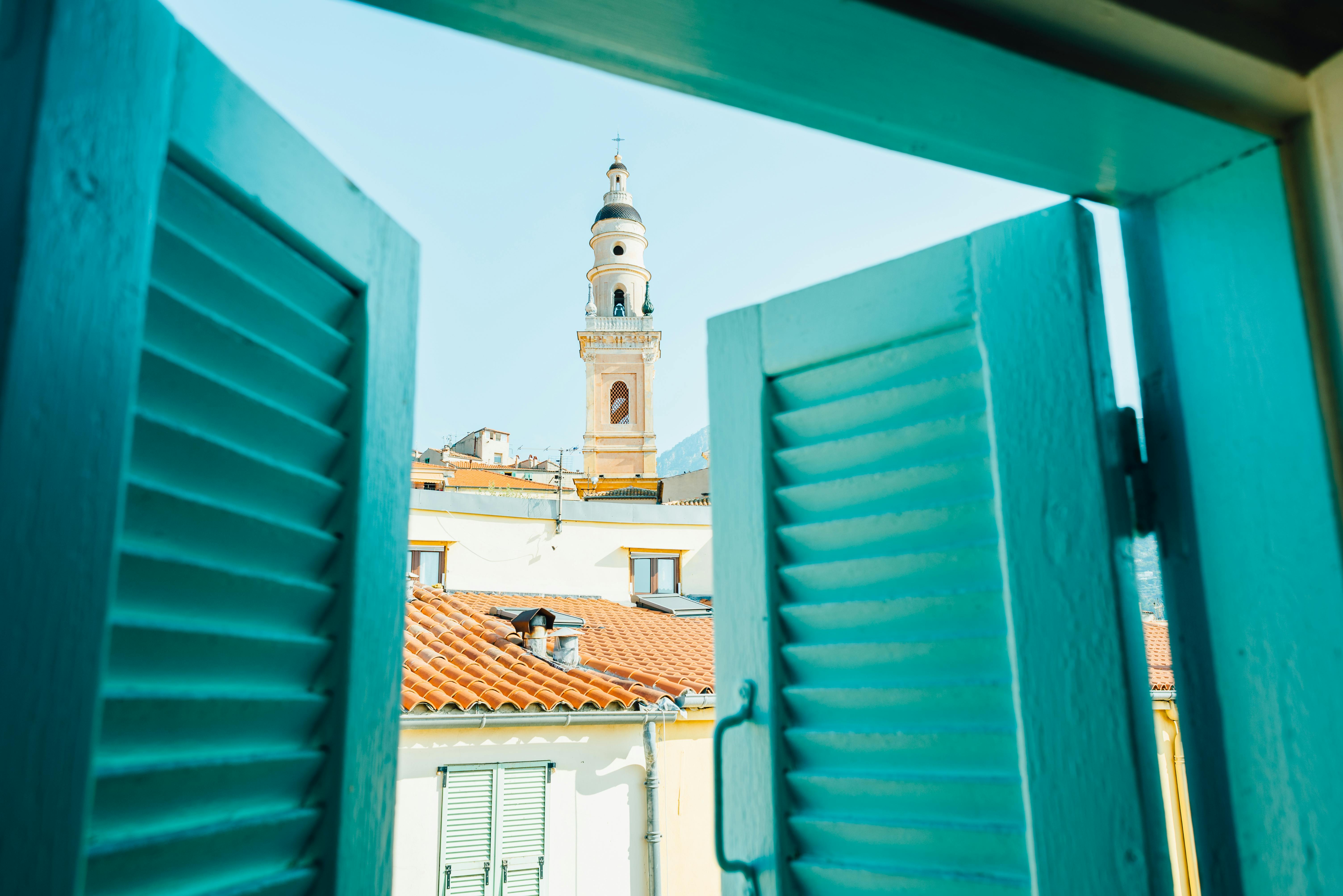 Vibrant view of church tower and rooftops through turquoise shutters, Mediterranean ambiance.