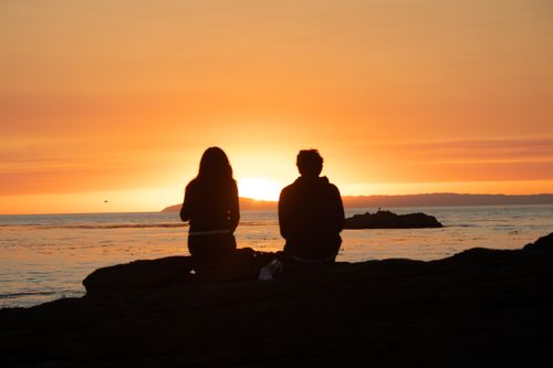 Free Couple sitting on rocks, gazing at the ocean during a vibrant sunset. Perfect for romantic themes. Stock Photo