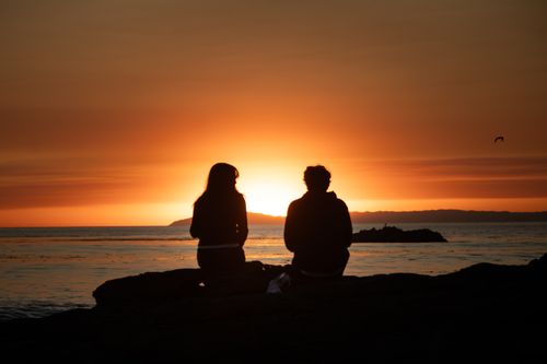 Free Silhouette of a couple sitting by the ocean enjoying a serene sunset view. Stock Photo