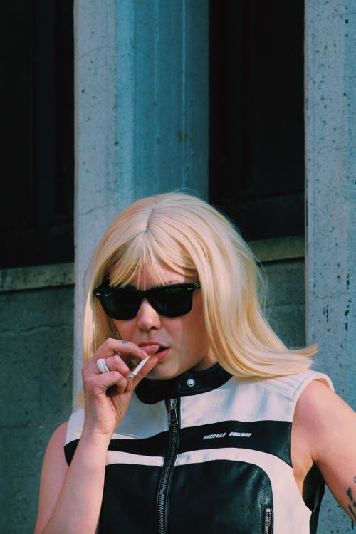 Free Stylish blonde woman in sunglasses smokes against an urban backdrop. Stock Photo