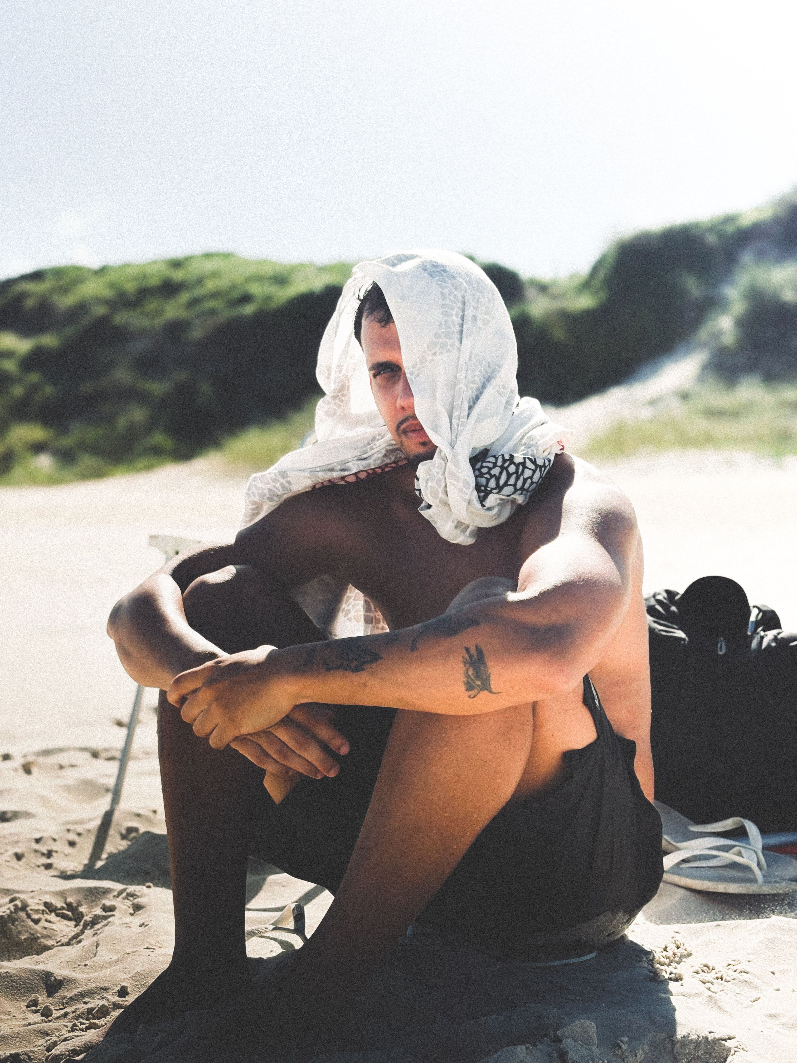 A young man sits casually on a beach, shaded by a scarf.