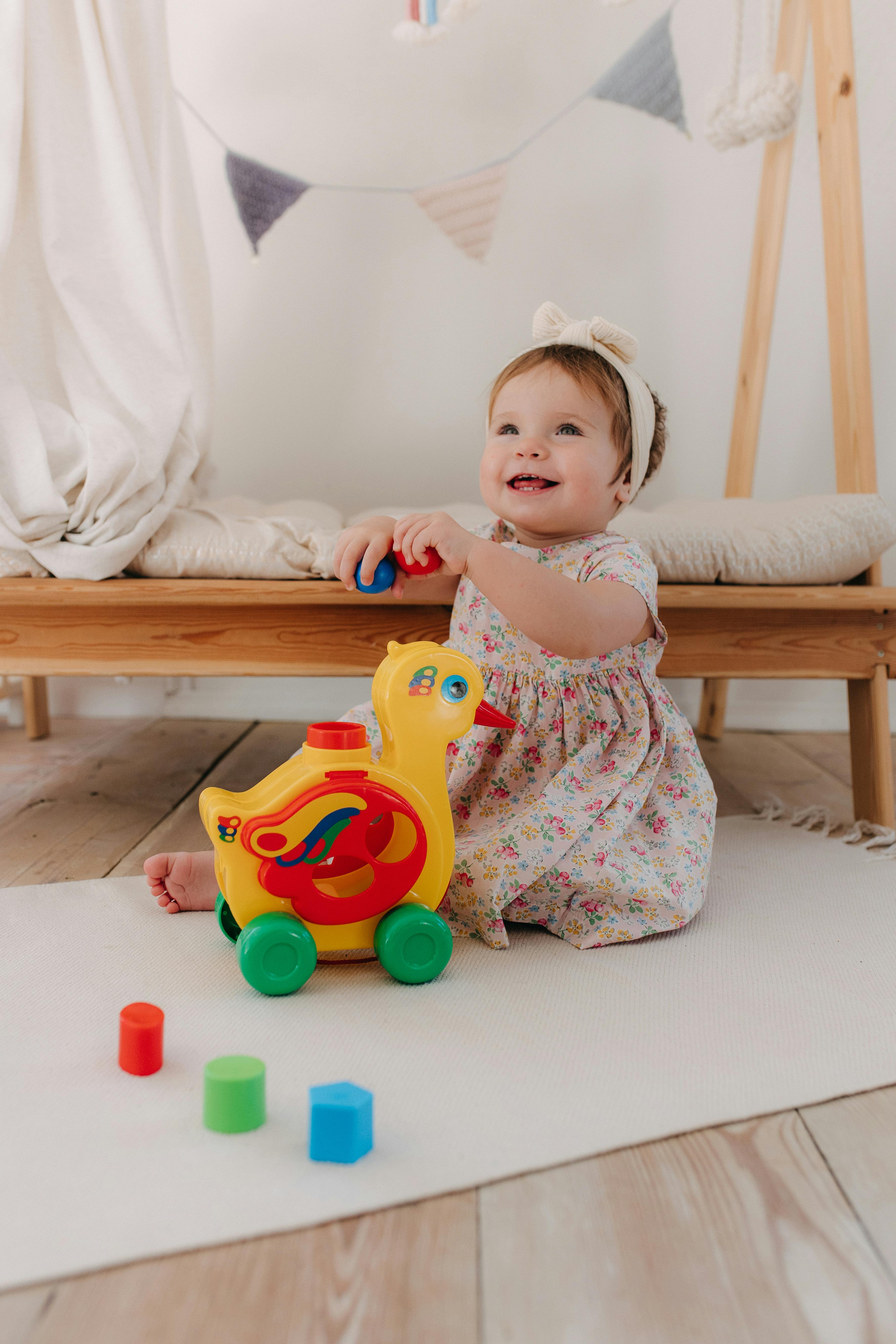 Adorable baby girl in floral dress playing with vibrant toy duck on the floor indoors.