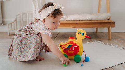 Cute baby girl playing with a colorful duck toy on a white rug in a cozy room.