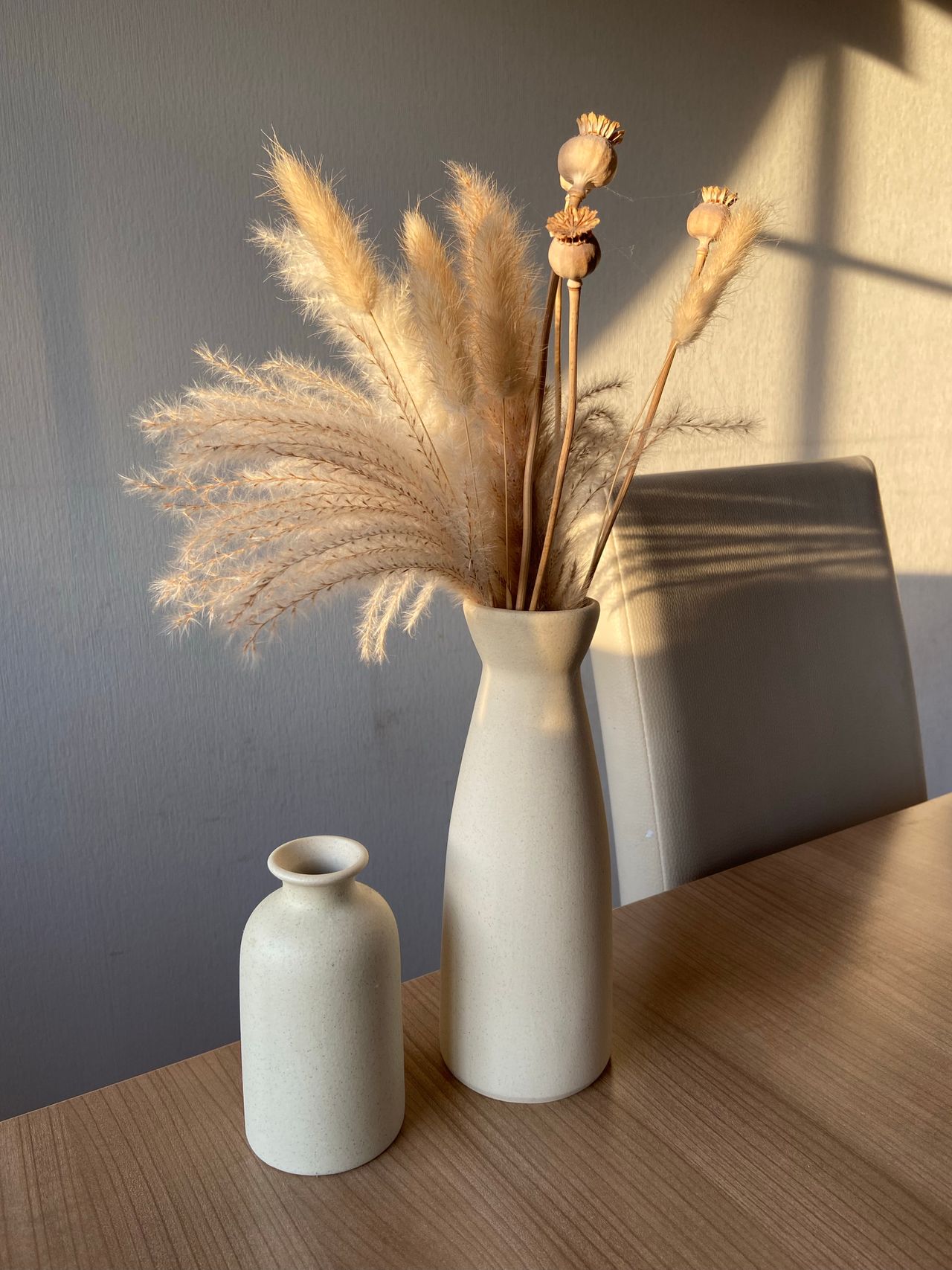 Dried pampas grass mixed with poppy seed heads in a tall cream vase on a dining table