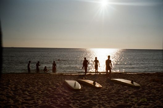 People silhouette, surfboards on beach, sunset reflecting off ocean waves.