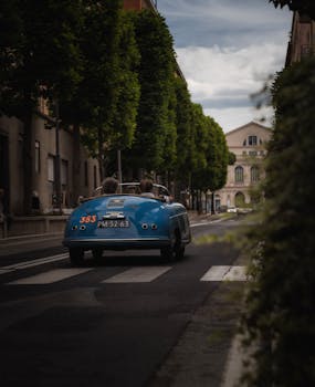 A classic blue car driving through a tree-lined city street, suggesting a vintage feel.