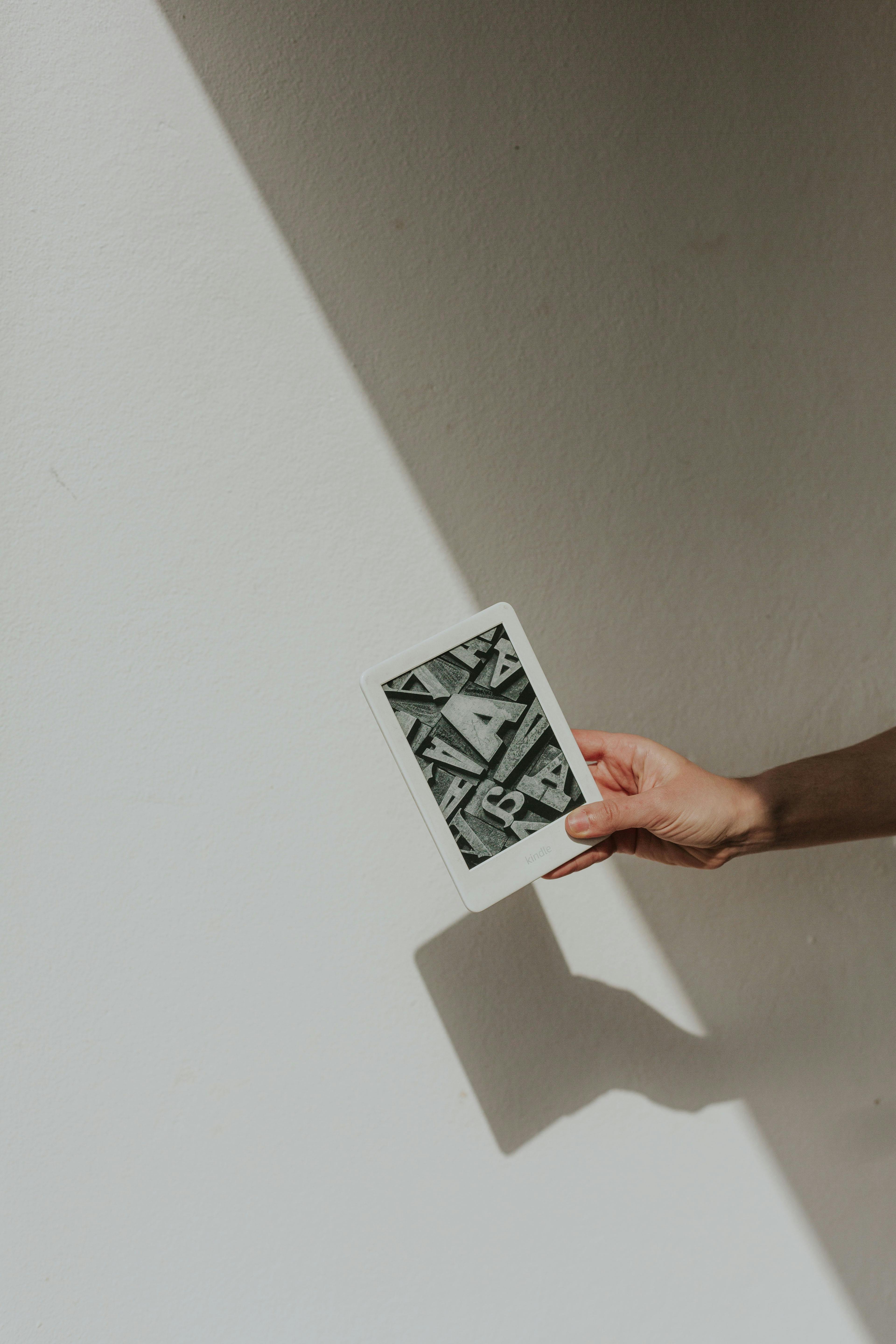 A minimalist photo featuring a hand holding a tablet against a light background, casting a shadow.