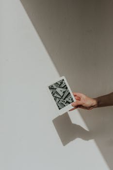 A minimalist photo featuring a hand holding a tablet against a light background, casting a shadow.