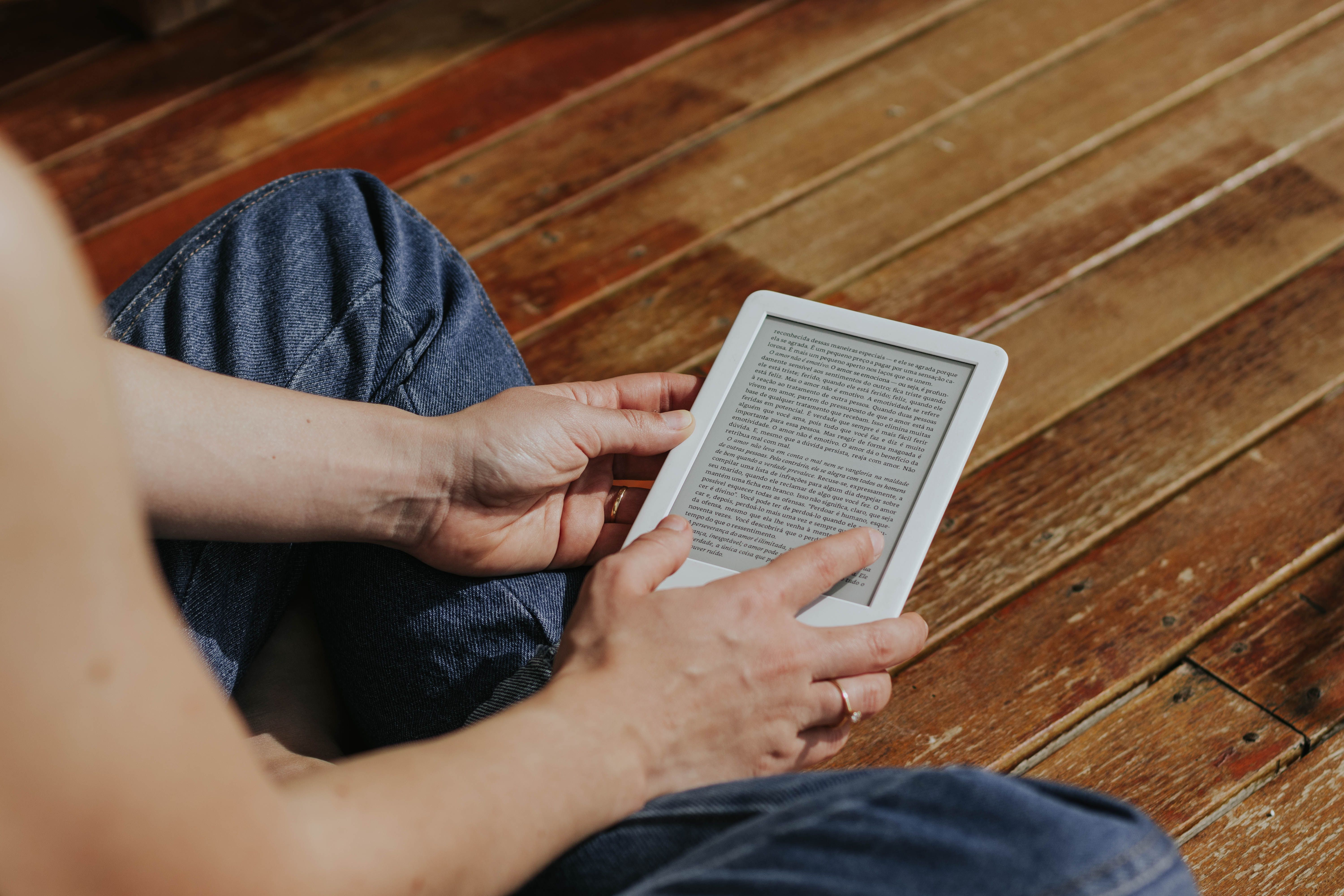 grátis Close de mãos segurando um leitor eletrônico em um ambiente interno, sobre um piso de madeira, enfatizando a leitura digital. Foto profissional