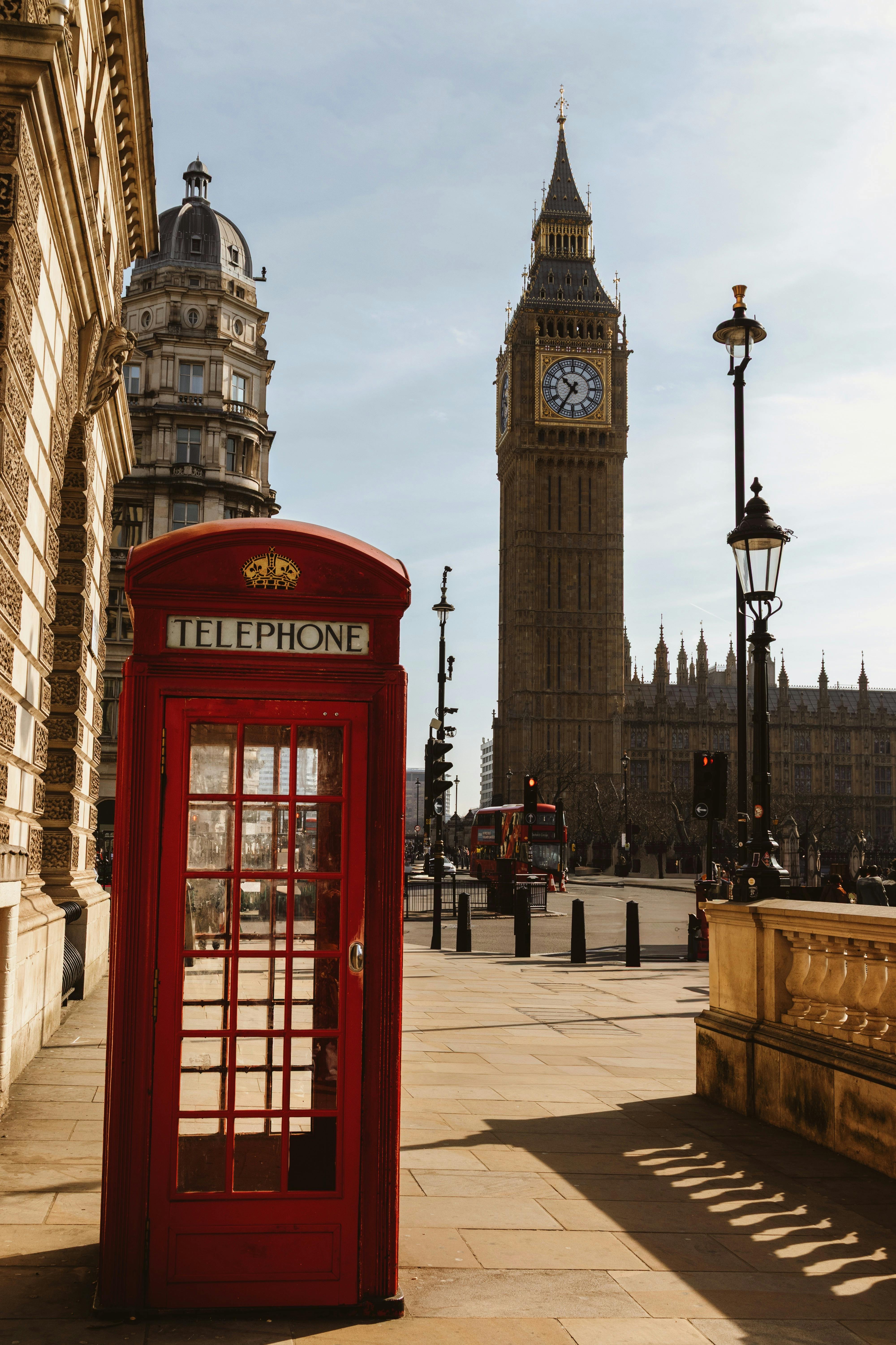 Red telephone booth with Big Ben and historic London architecture in daylight.