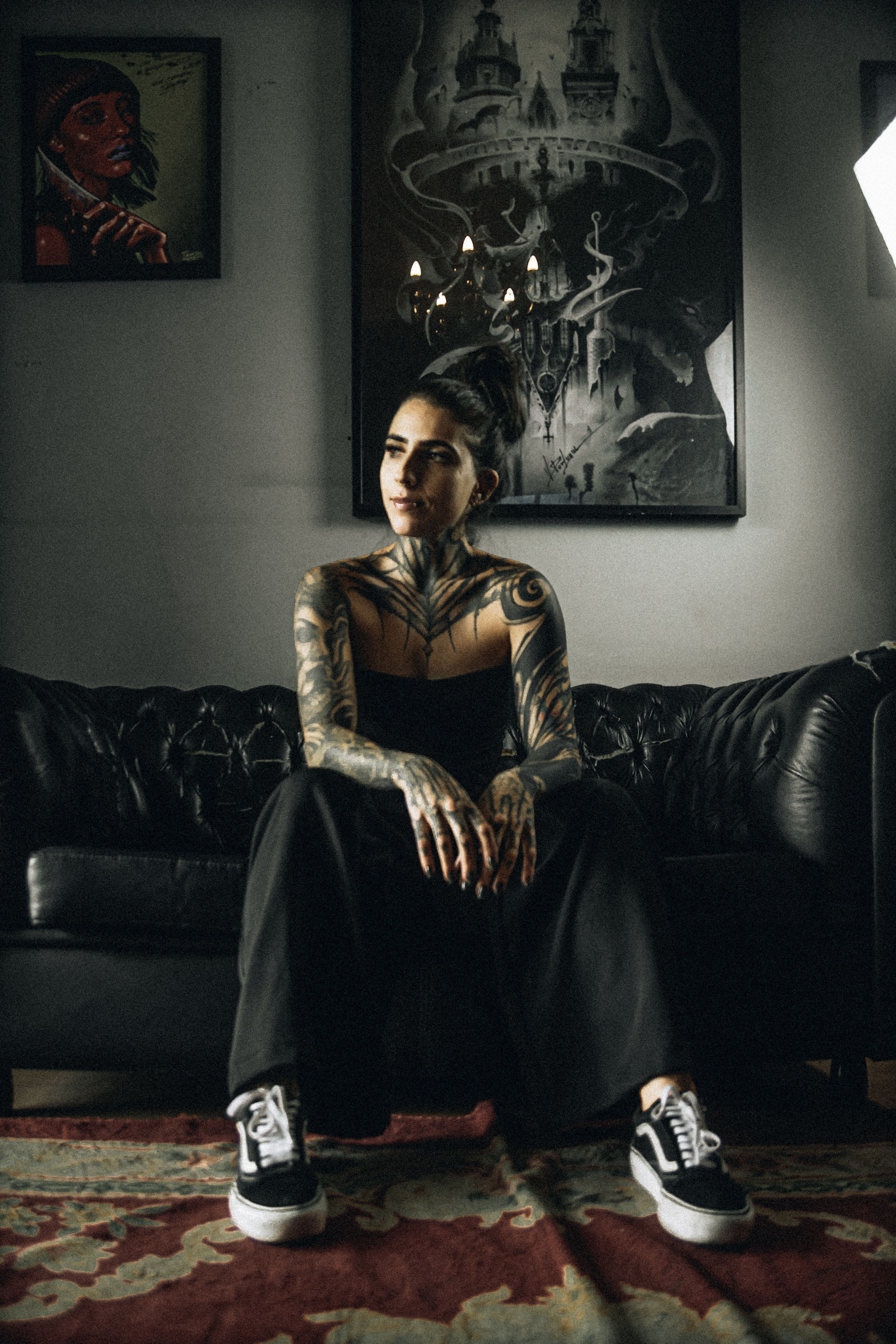 Tattooed woman in black outfit sitting on leather sofa in artistic setting.