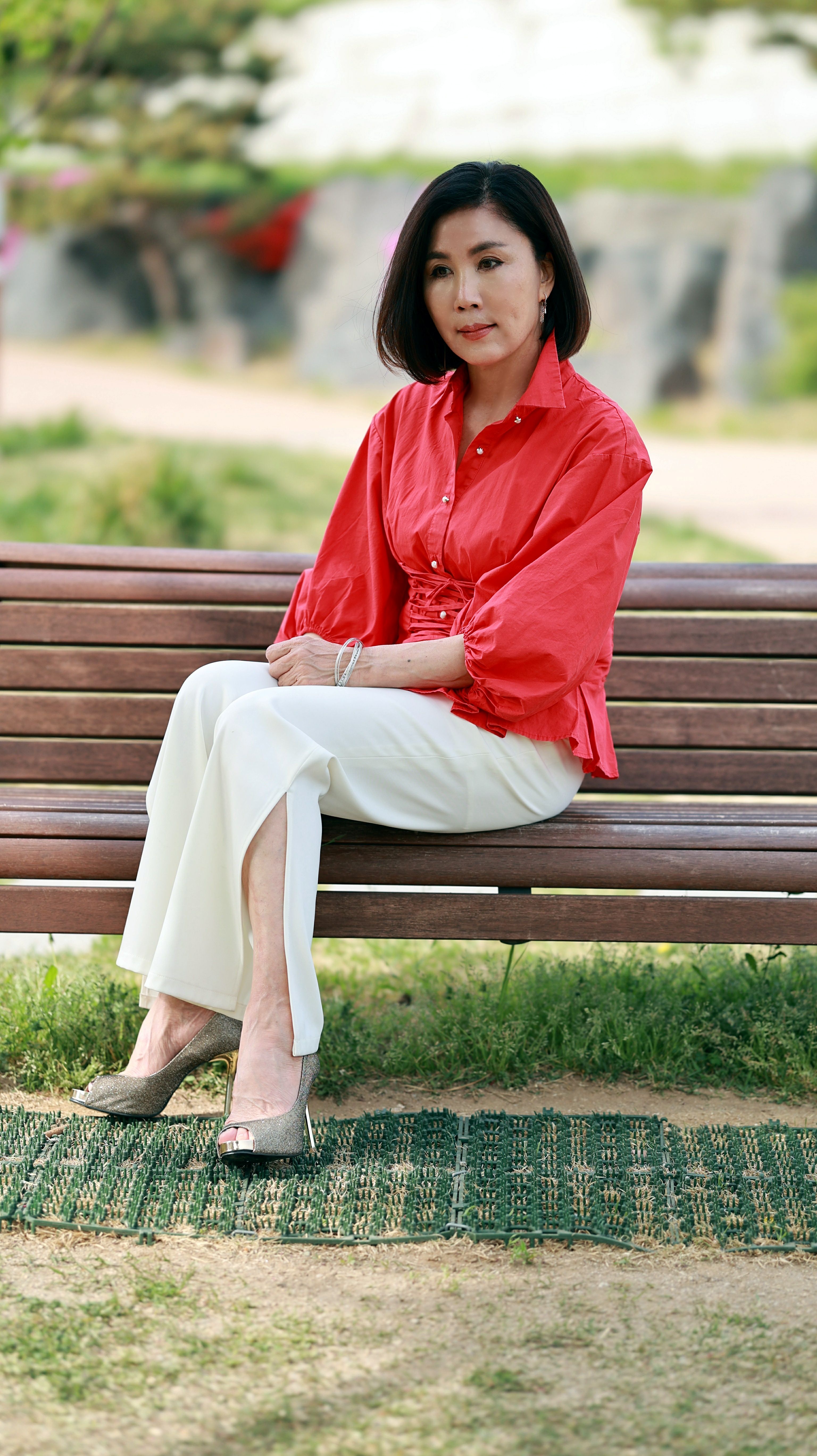Stylish woman in a red blouse sitting on a bench, Seoul park scene, outdoors on a sunny day.
