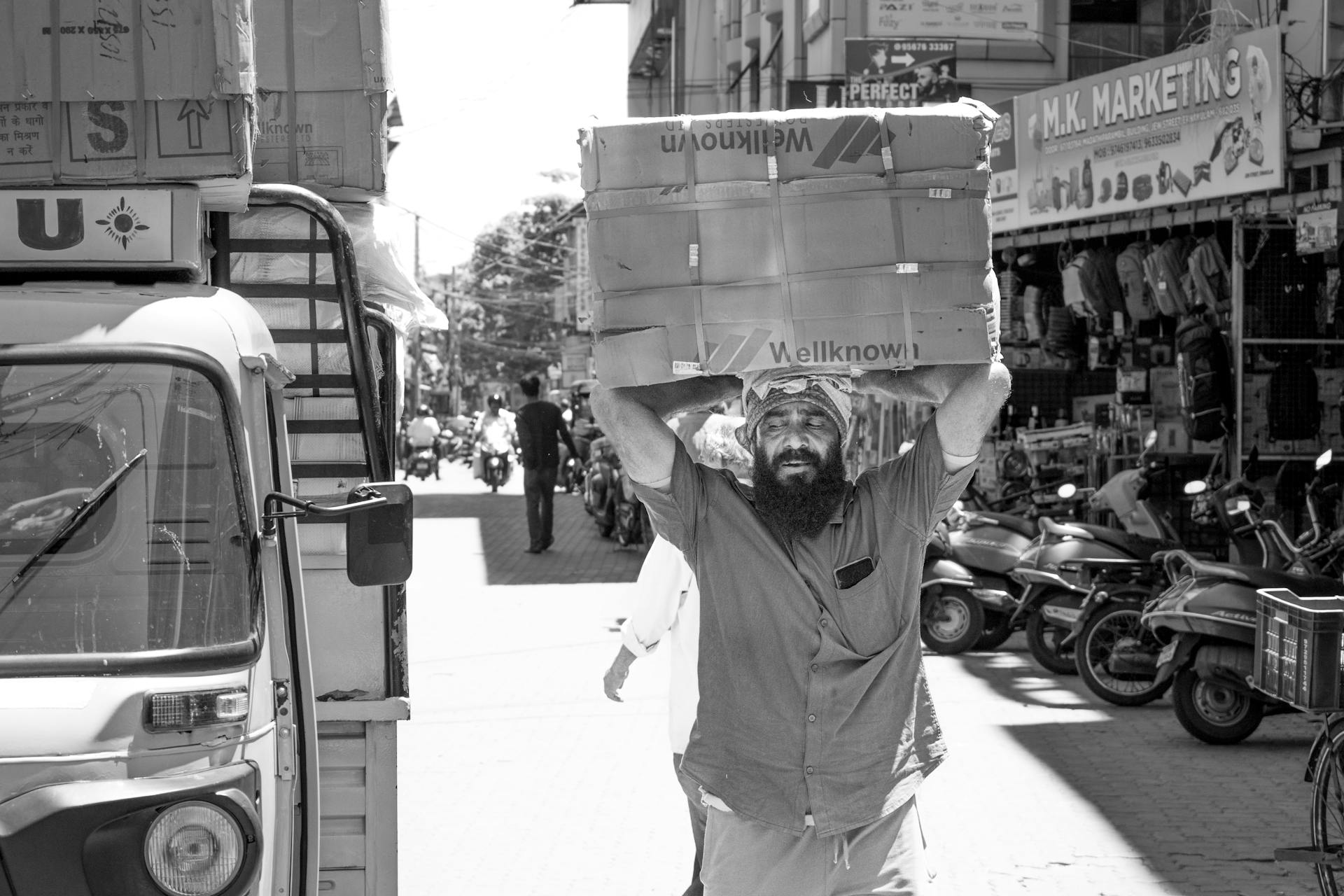 A bearded man carries boxes on his head in a bustling Kochi market, showcasing the daily life hustle.