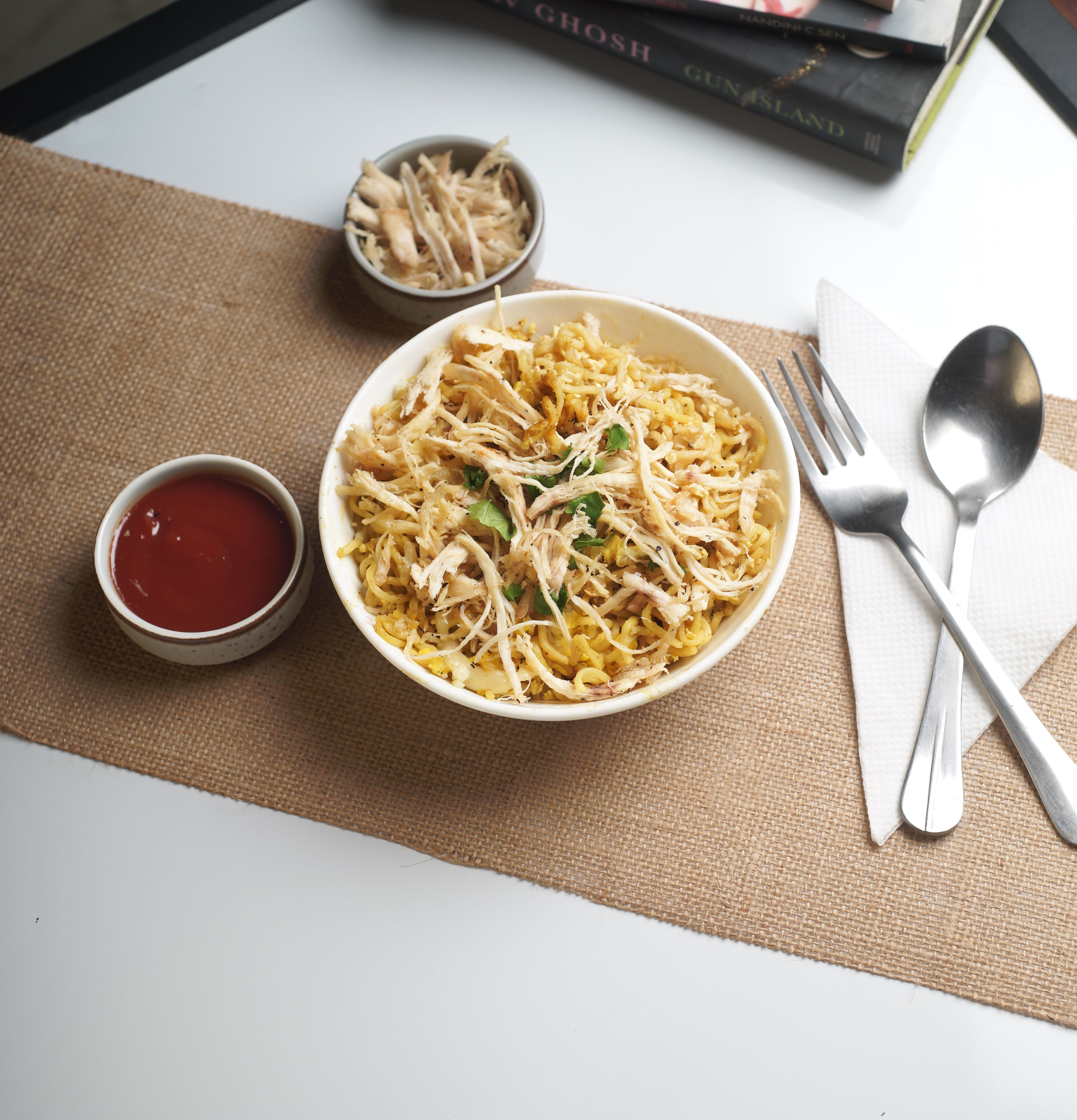 Delicious bowl of chicken noodles with tomato sauce, served on a burlap mat with utensils on a white table.