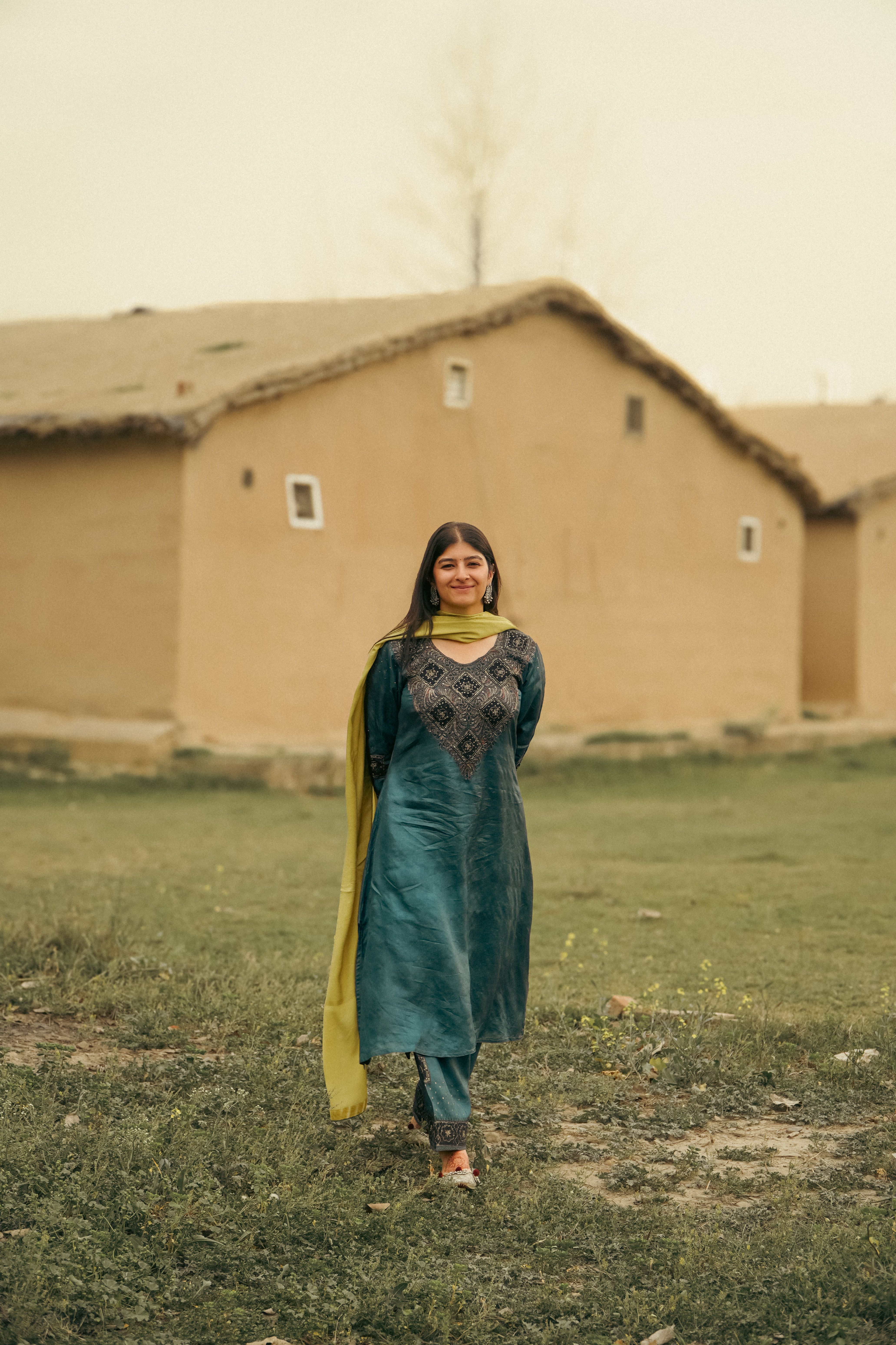 A woman in a vibrant traditional outfit stands outside rustic houses.
