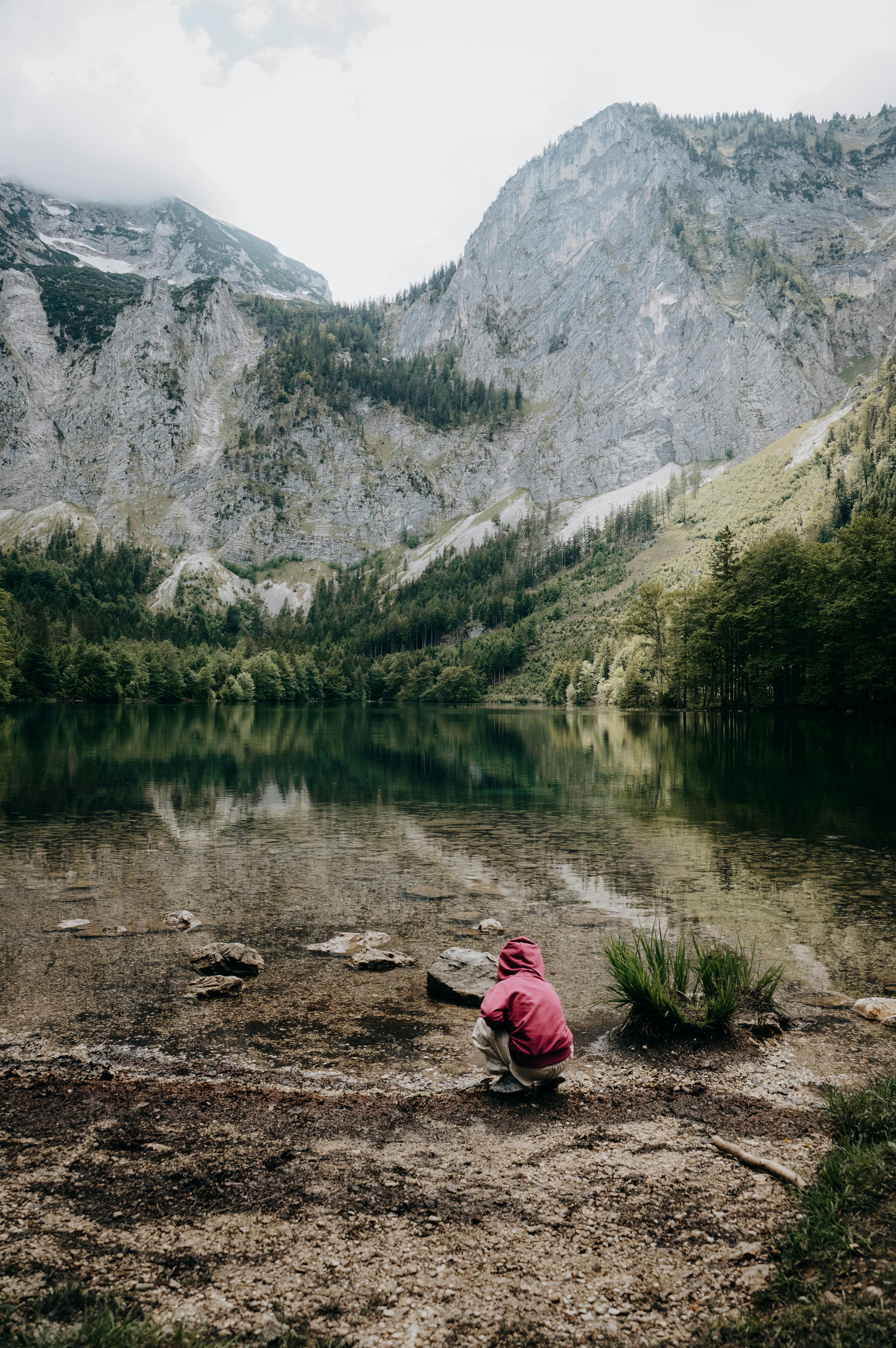 A child in a red hoodie explores the tranquil shore of a mountain lake, with towering cliffs in the background.