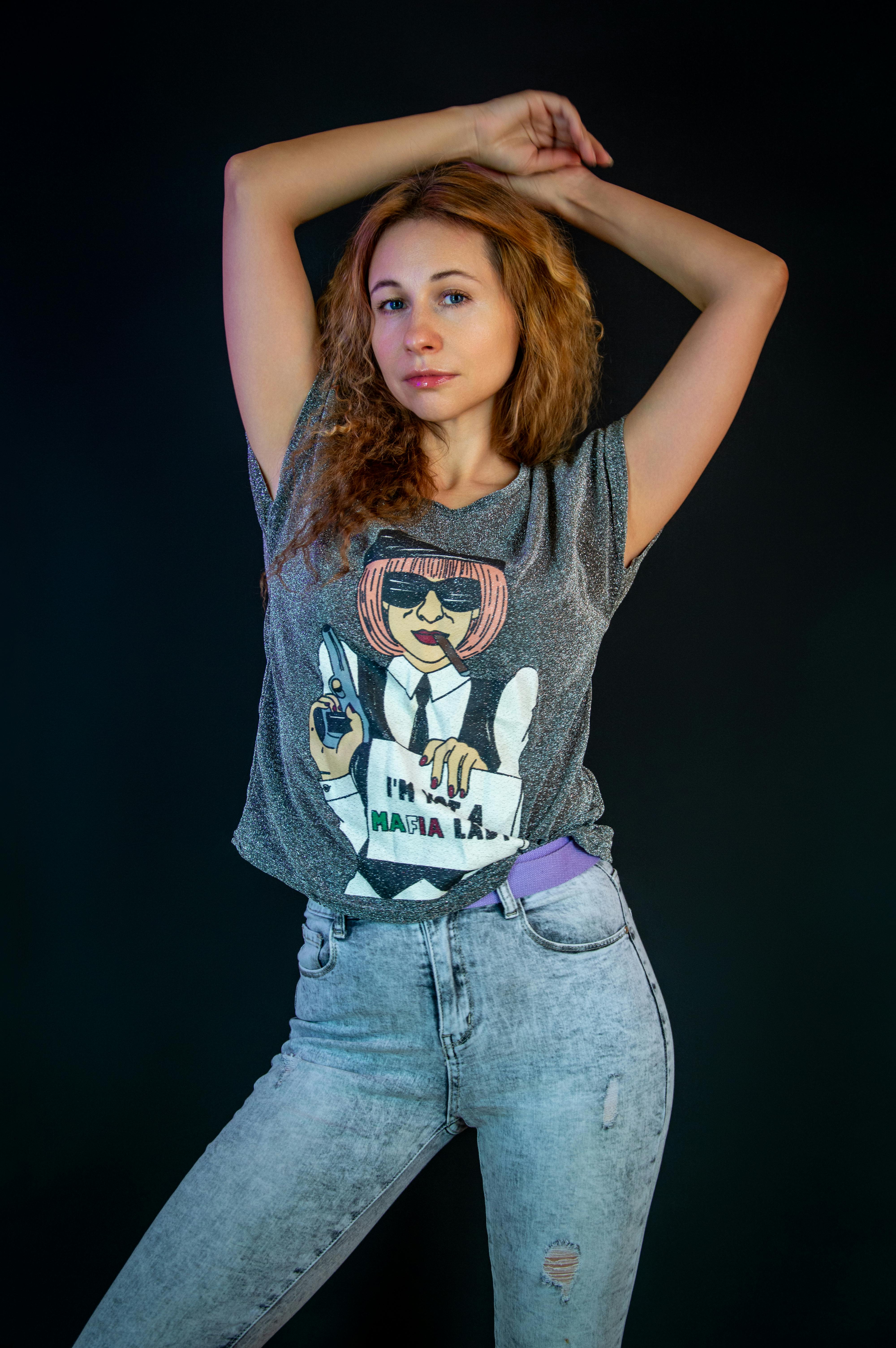 Woman striking a confident pose in gray tee and jeans against a black background.