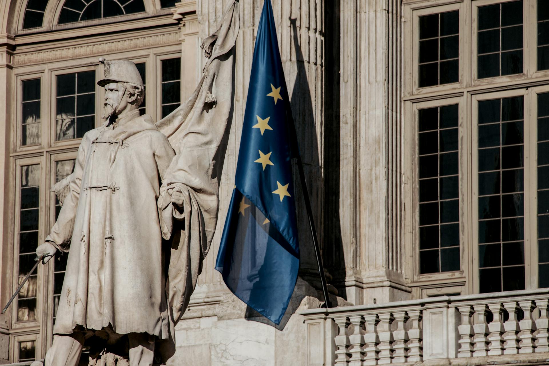 European Union flag waving beside a historic statue on stone building facade.