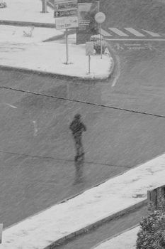 Black and white image of a person walking during snowfall on an urban road.