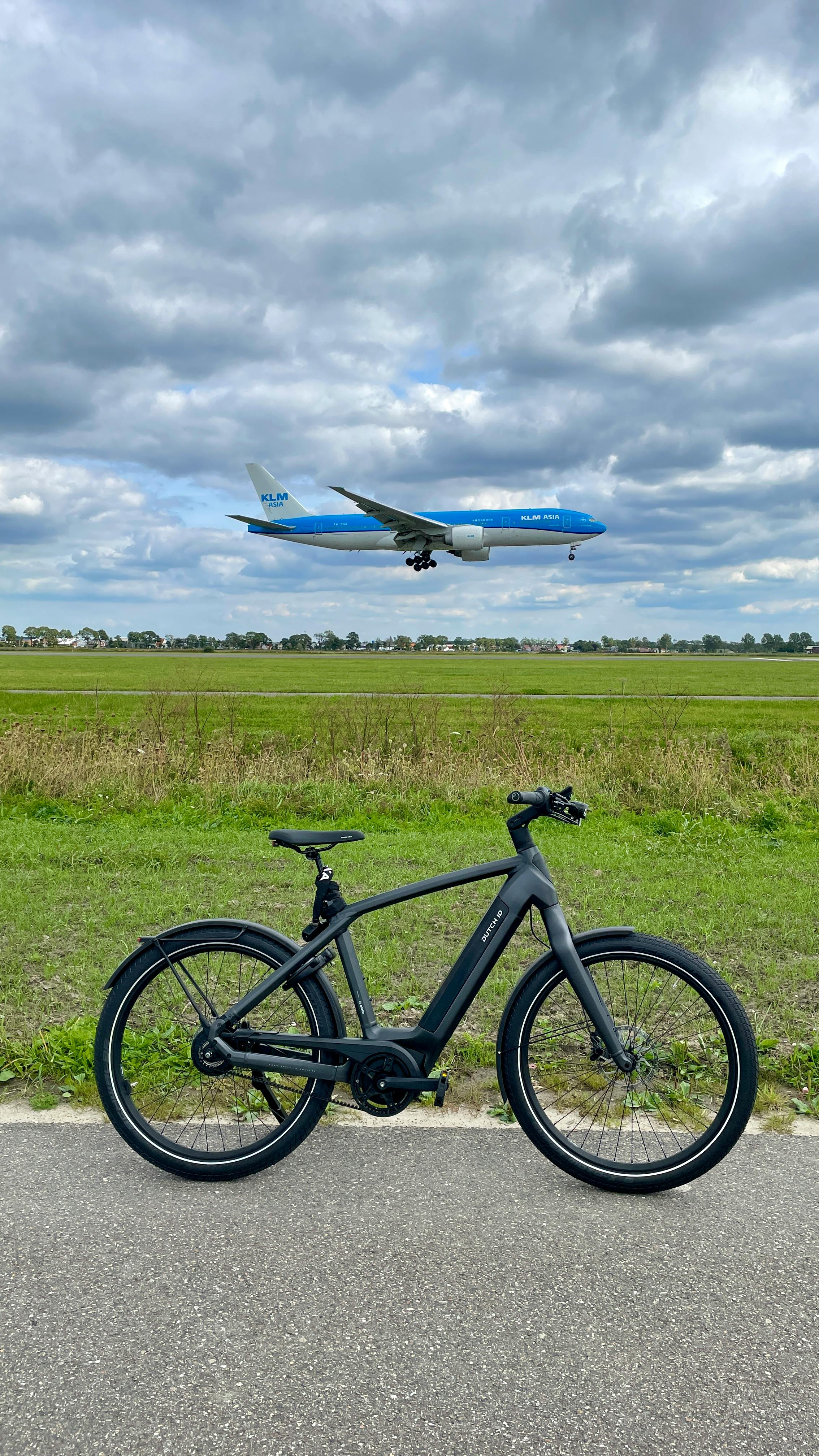 A black bicycle parked on a path with a plane flying overhead against a cloudy sky.