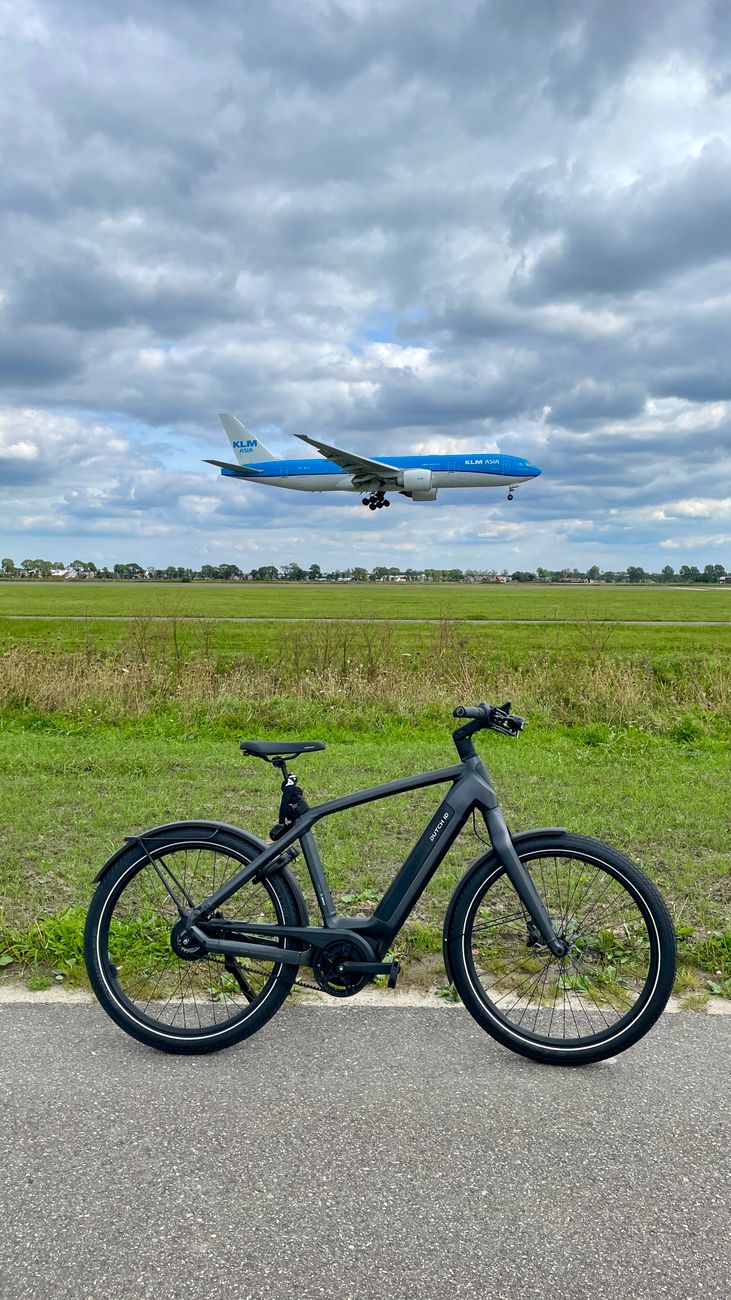 Een zwarte fiets geparkeerd op een pad met een vliegtuig dat boven het hoofd vliegt tegen een bewolkte lucht.