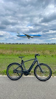 A black bicycle parked on a path with a plane flying overhead against a cloudy sky.