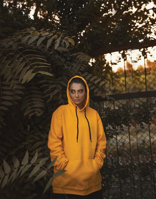 Free Woman in yellow hoodie standing next to foliage during sunset, posing for a fashion shoot. Stock Photo