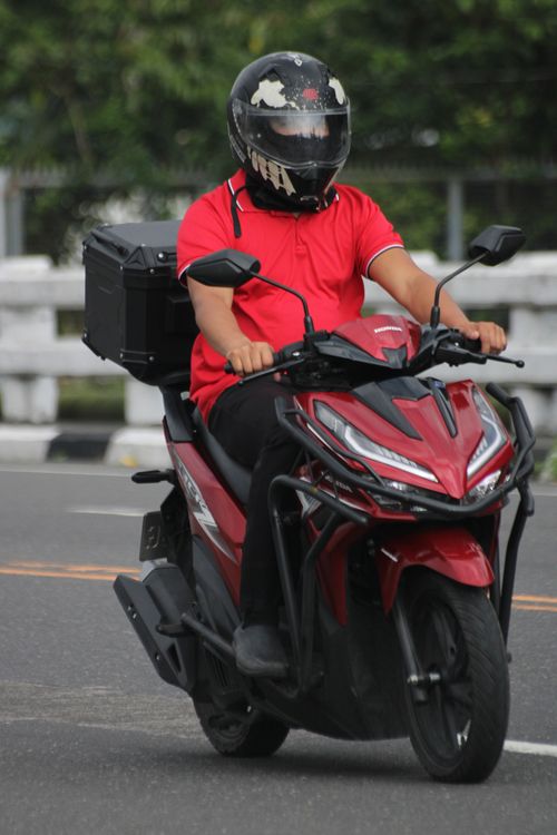 Free A motorcyclist in red delivery uniform riding a scooter on a city street. Stock Photo