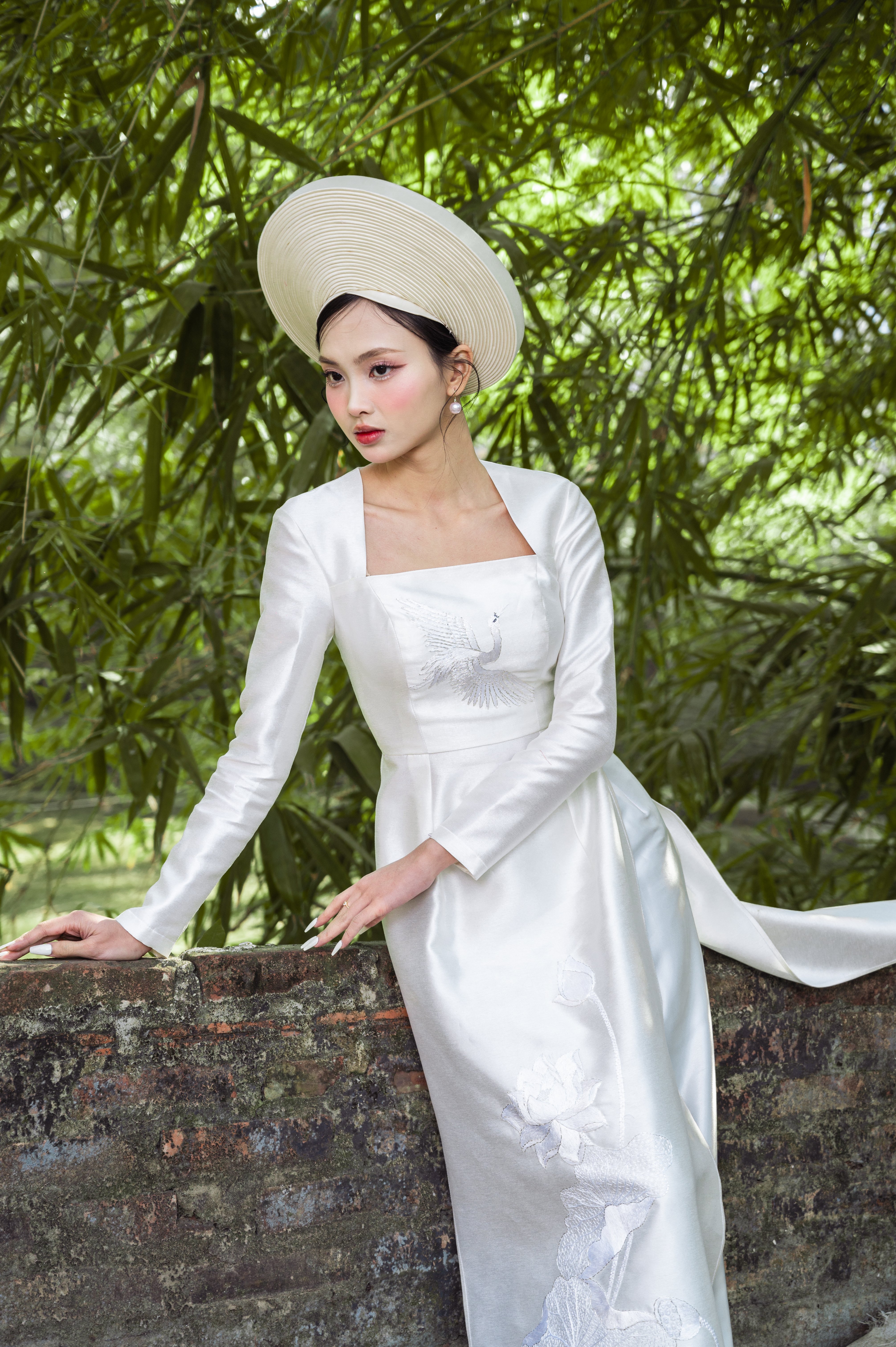 A woman stands by a wall in a white ao dai with a traditional hat.