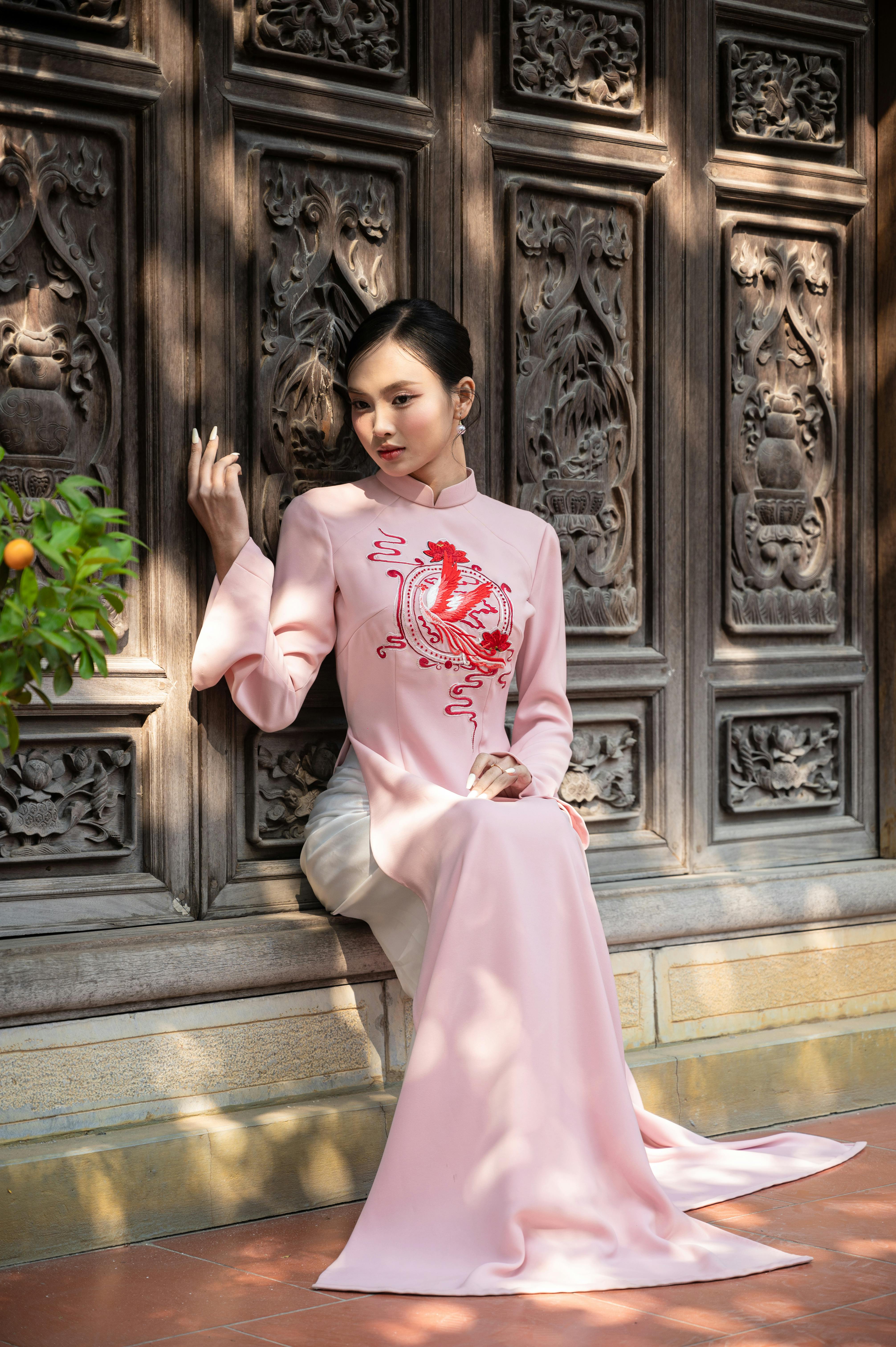 Woman in a pink áo dài sitting by ornate wooden door under soft sunlight.