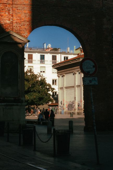 A city corner in Milan captured through an arched brick frame with warm sunlight casting shadows.