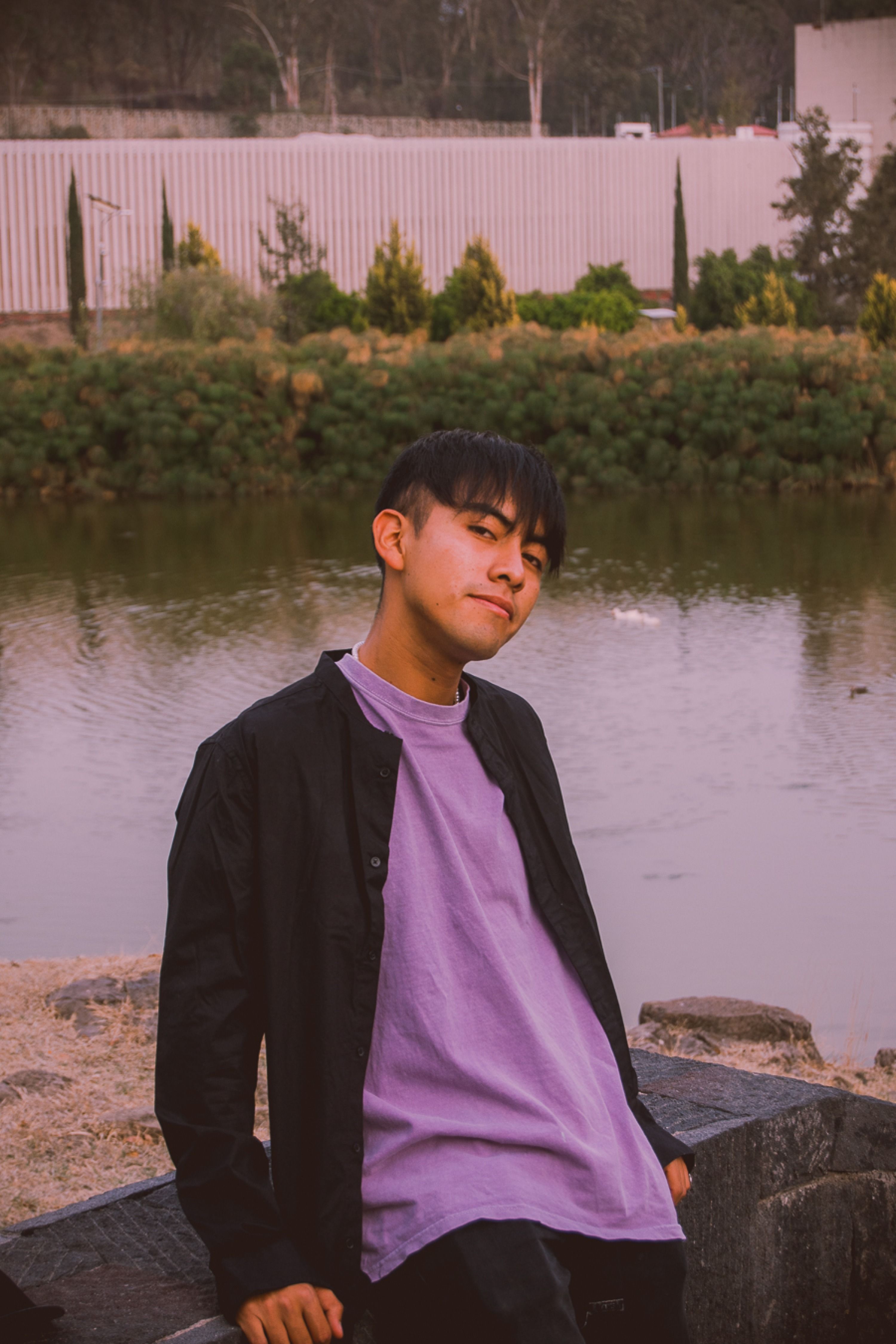 A young man in a purple shirt and black jacket by the riverside, exuding a relaxed vibe.